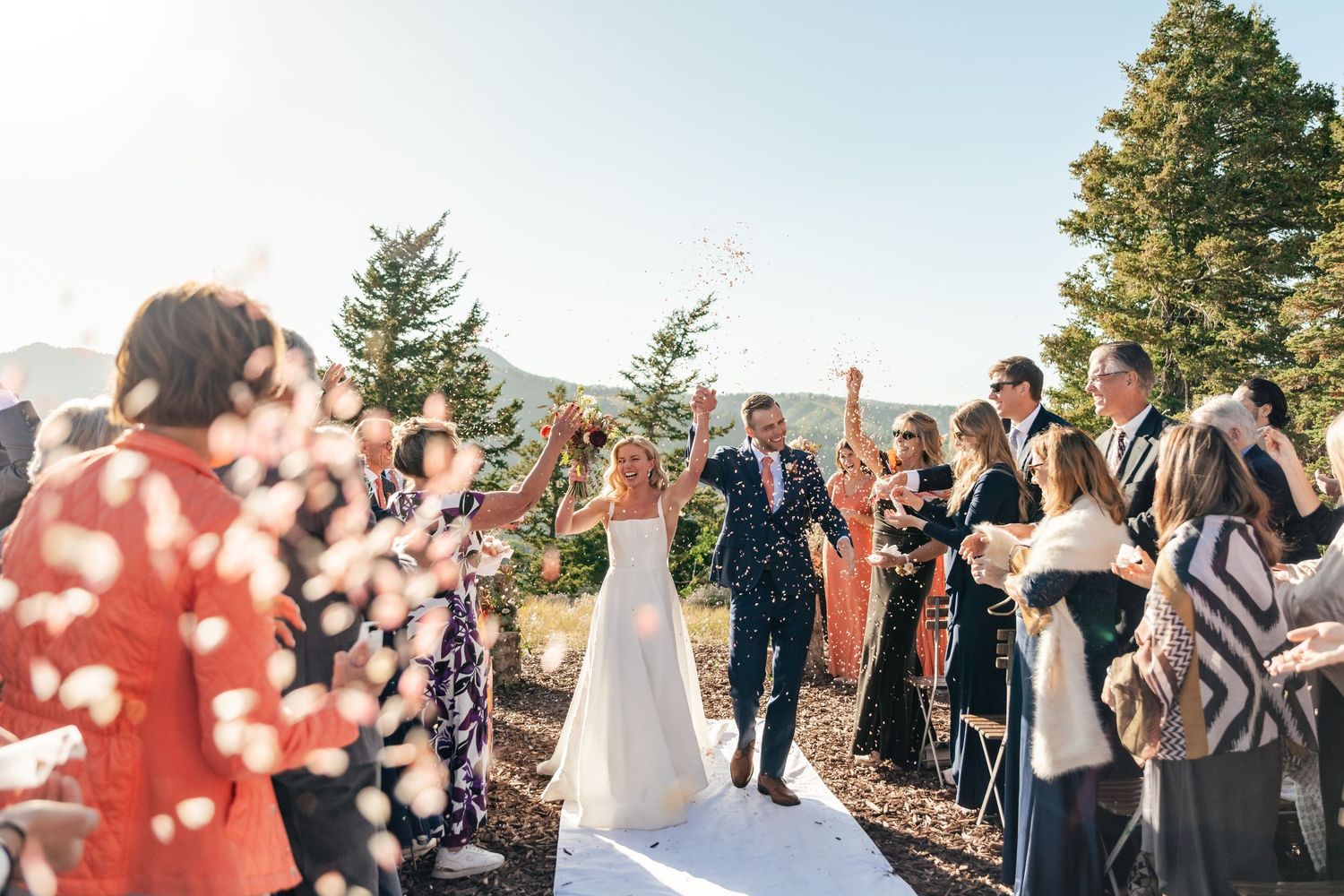 Wedding guests celebrate and throw confetti as newlyweds walk down the aisle at an outdoor mountain ceremony. Park City Wedding Photographer