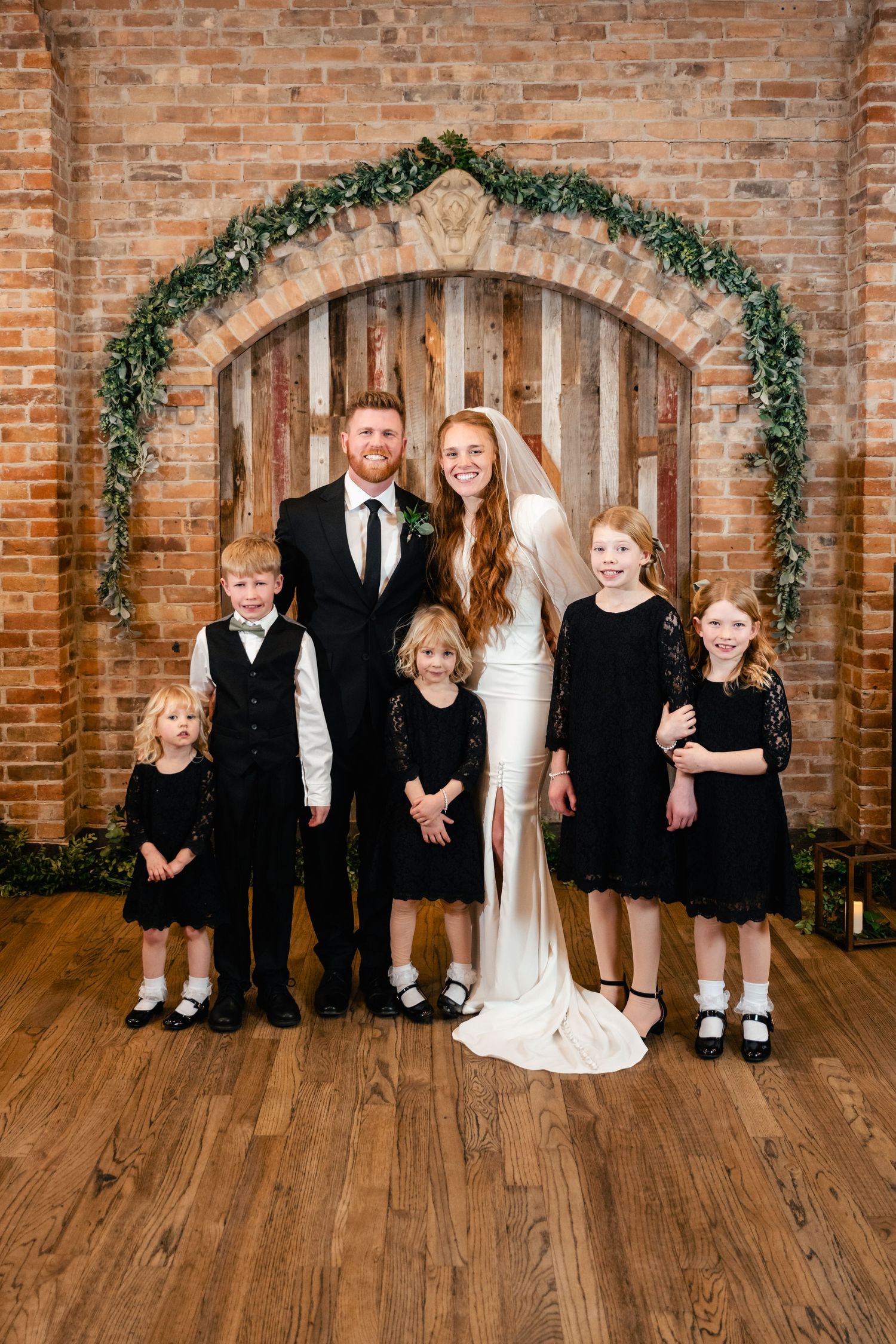 Wedding party posing under a greenery arch against exposed brick wall backdrop.