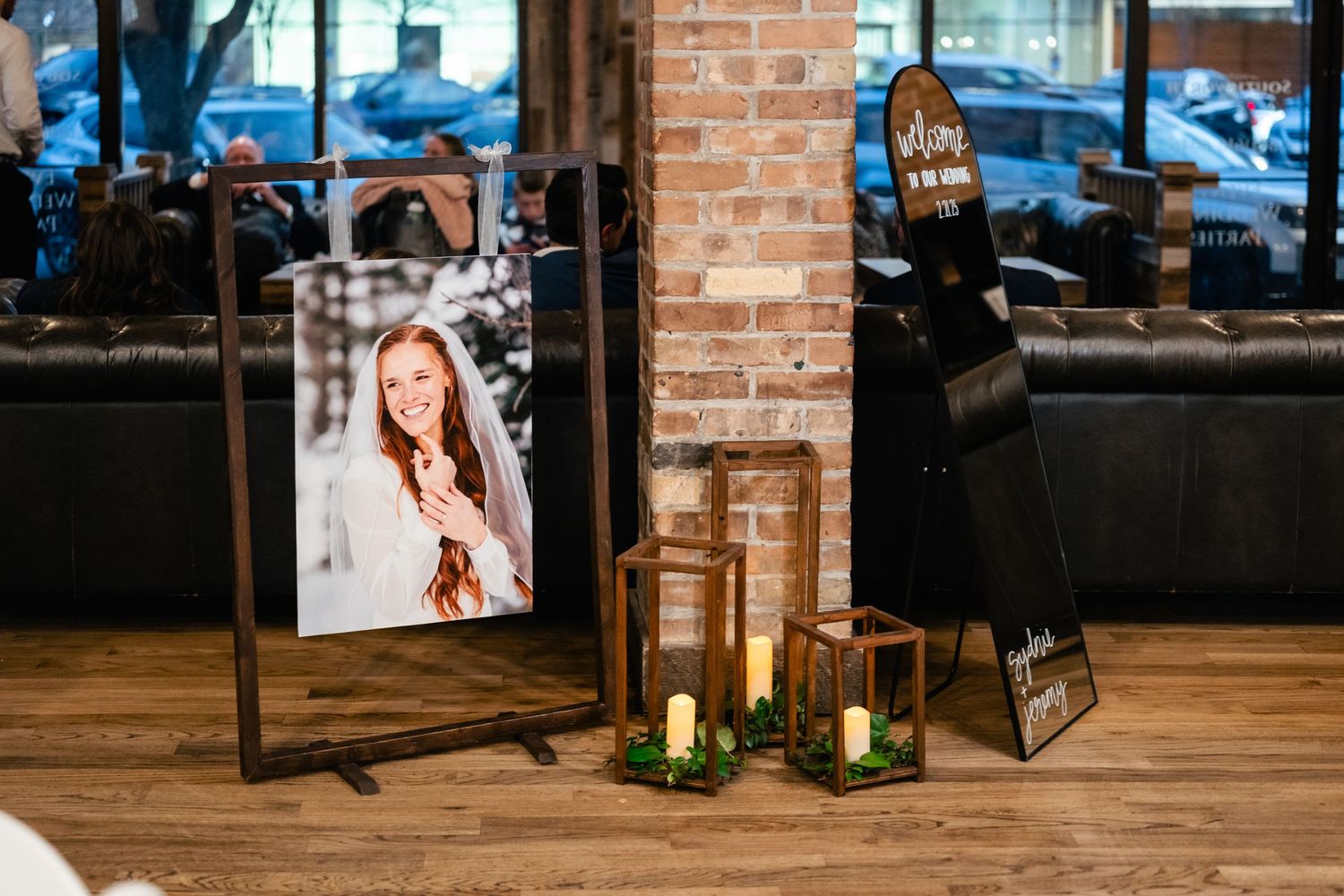 A framed portrait sits on a wooden floor beside glowing candle lanterns near an exposed brick wall.