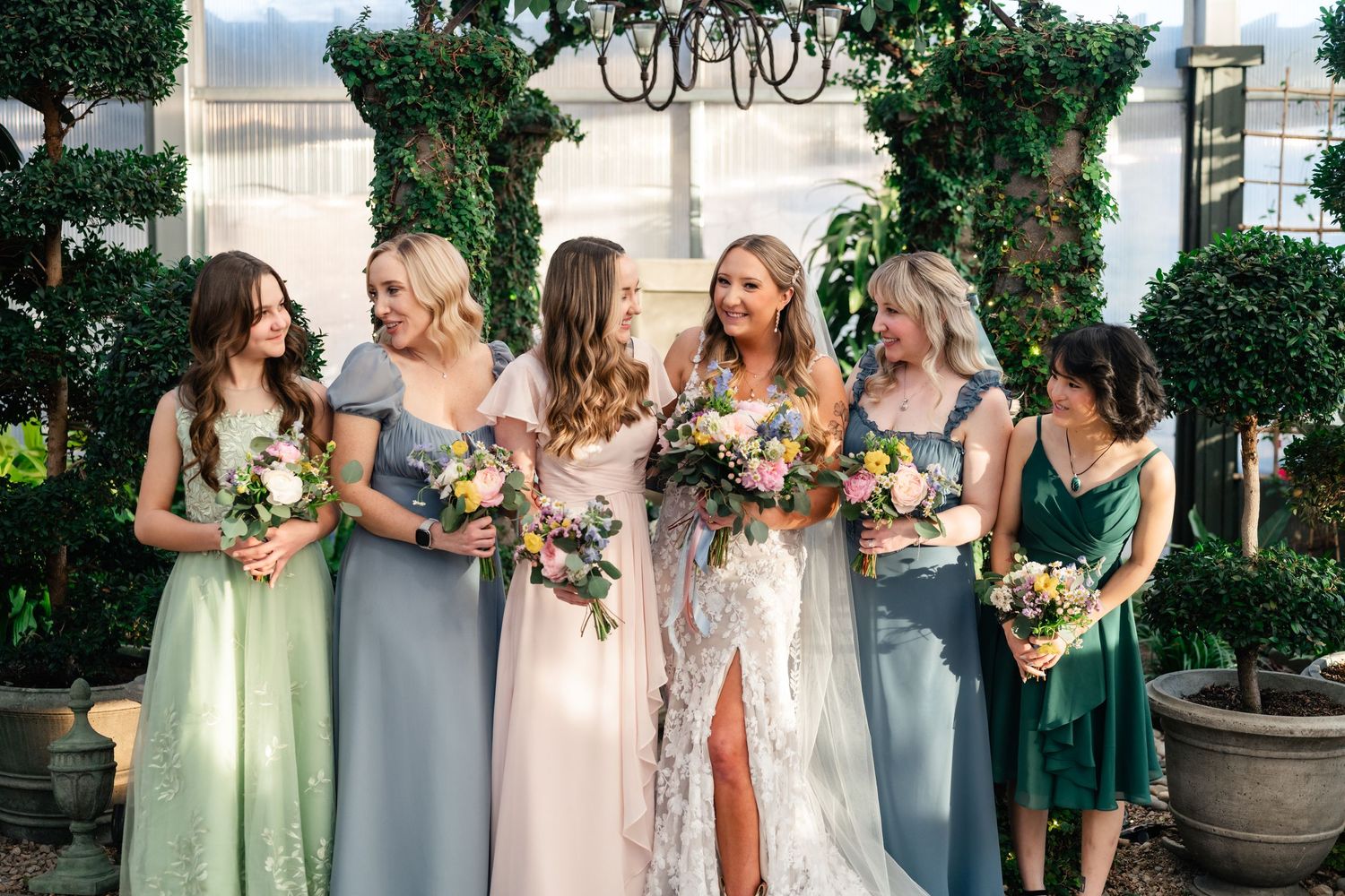 Bride and bridesmaids holding floral bouquets smile together in front of greenery-covered greenhouse backdrop.