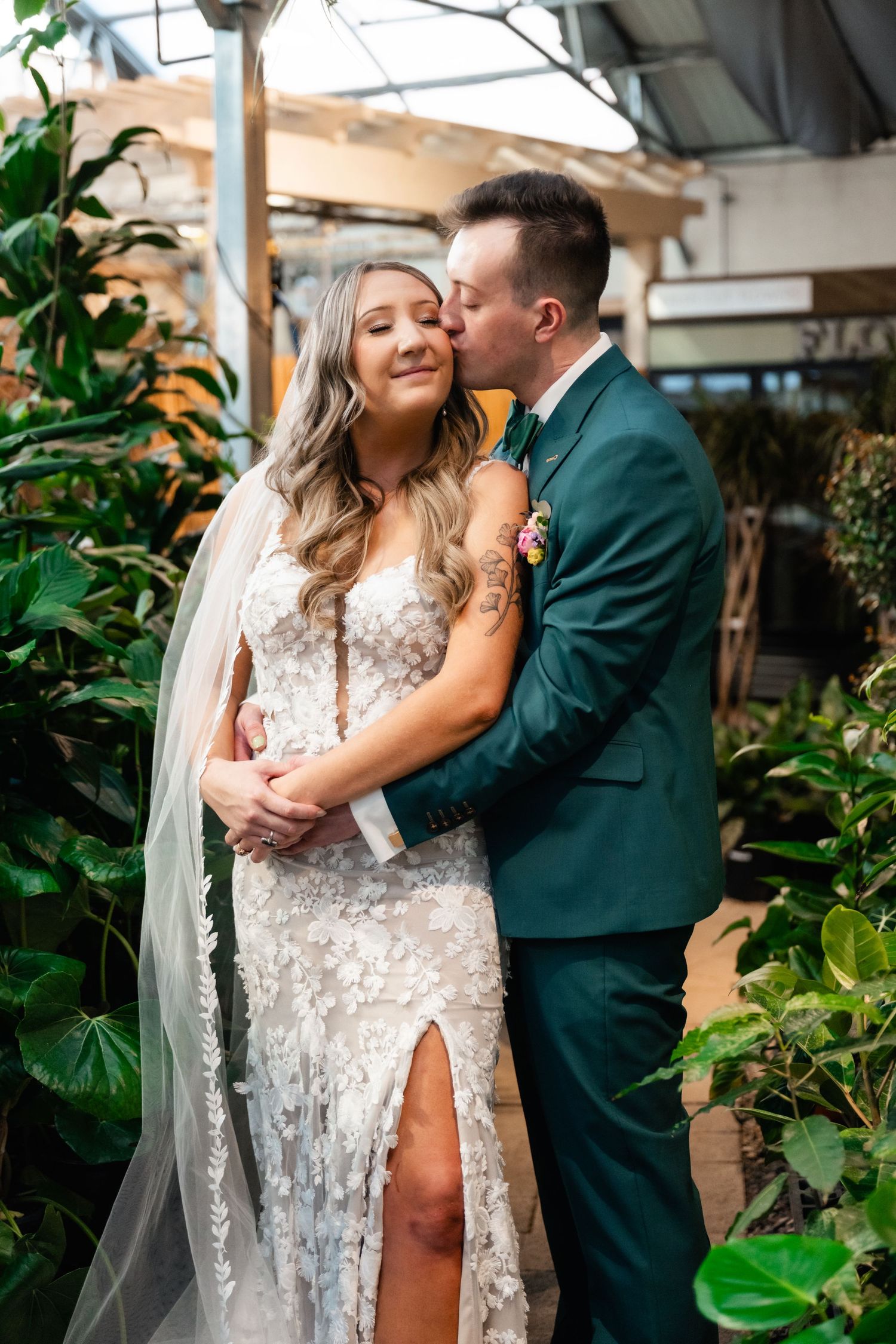 Couple in formal wedding attire embracing in front of lush greenery at an outdoor venue.