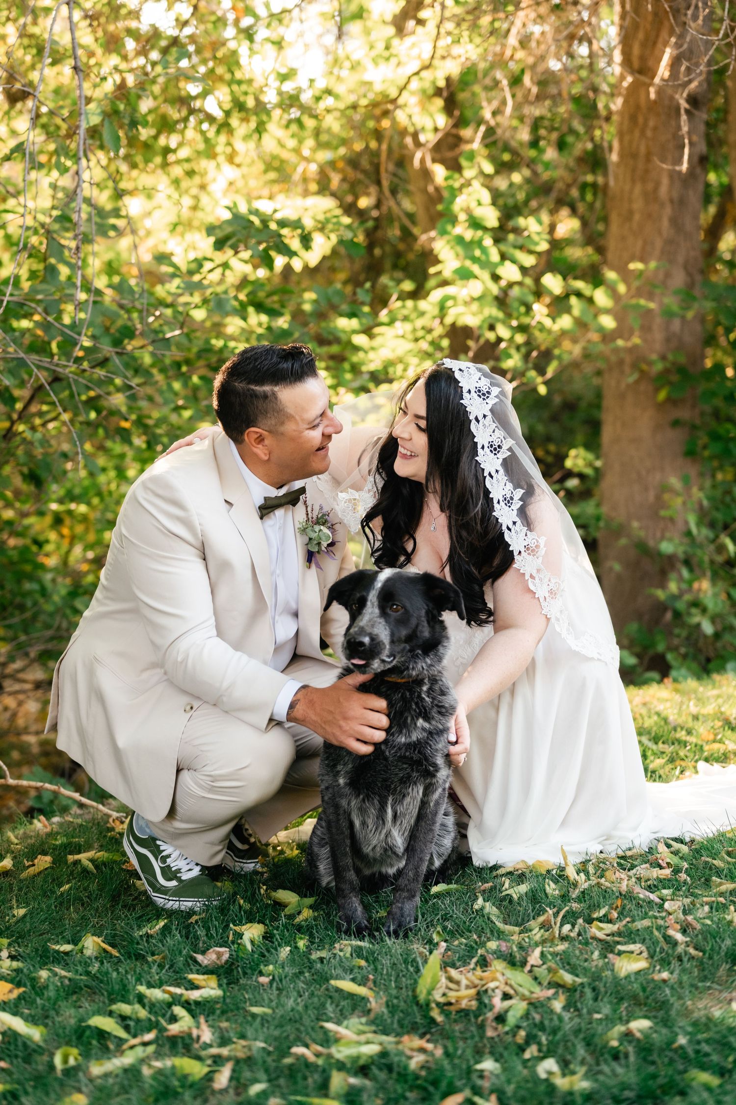 A newlywed couple crouches with their black dog on lush grass in a garden setting.