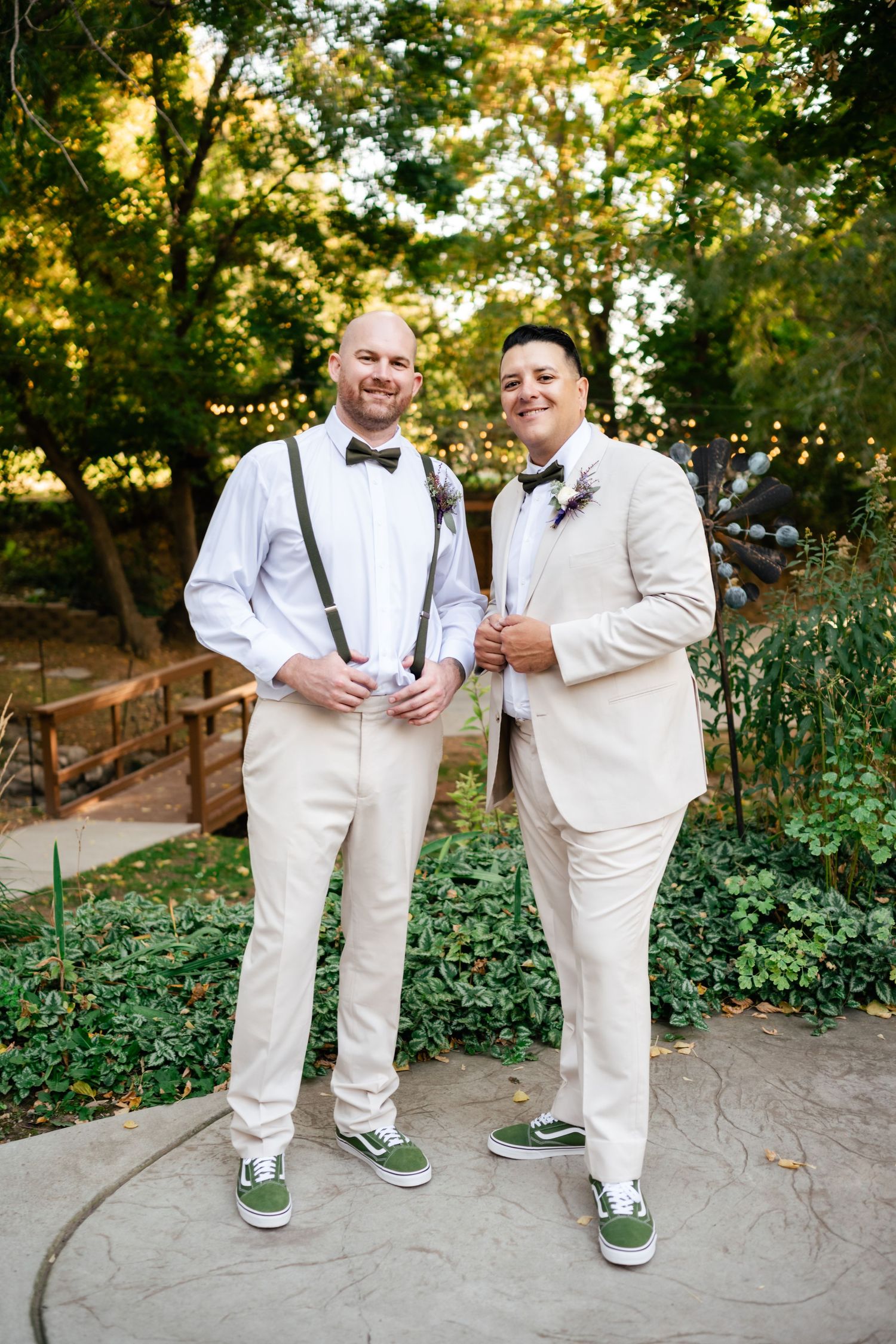 A couple wearing matching cream-colored suits with green sneakers pose together at an outdoor garden wedding ceremony.