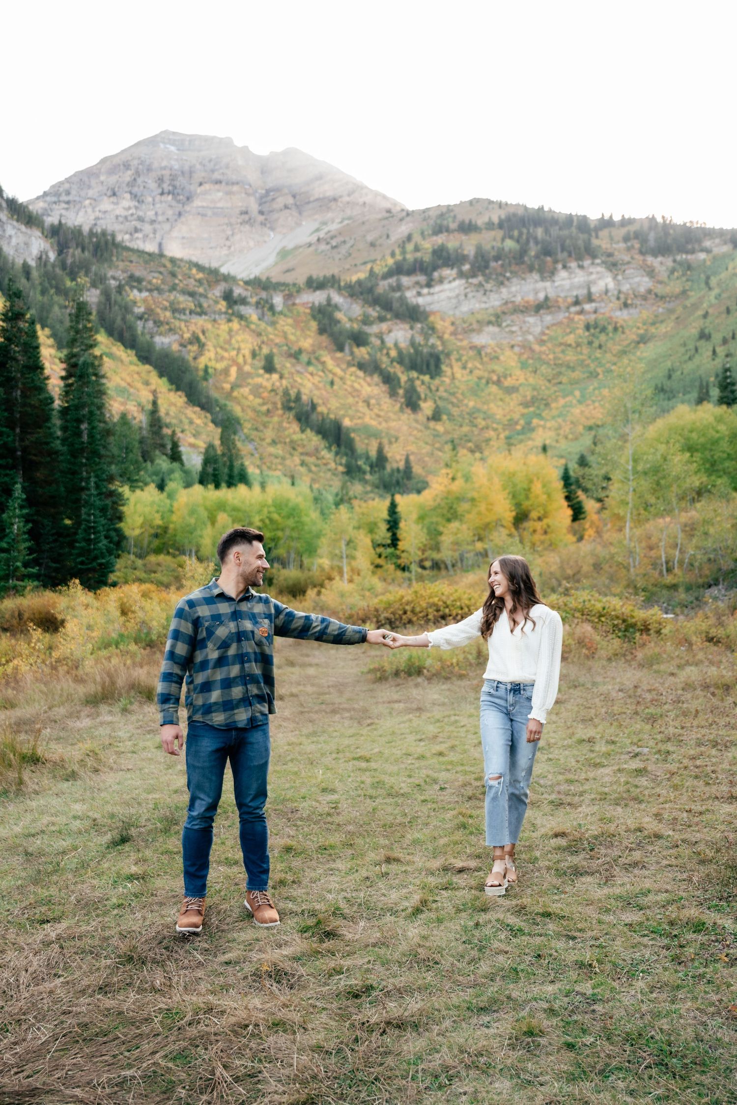 A couple holds hands in a scenic mountain meadow surrounded by golden fall foliage and evergreen trees.