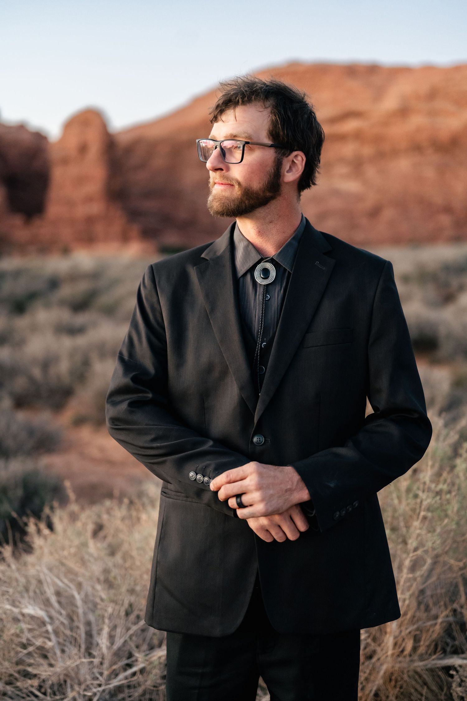 Man in black formal suit and bolo tie poses against red desert rock formations.