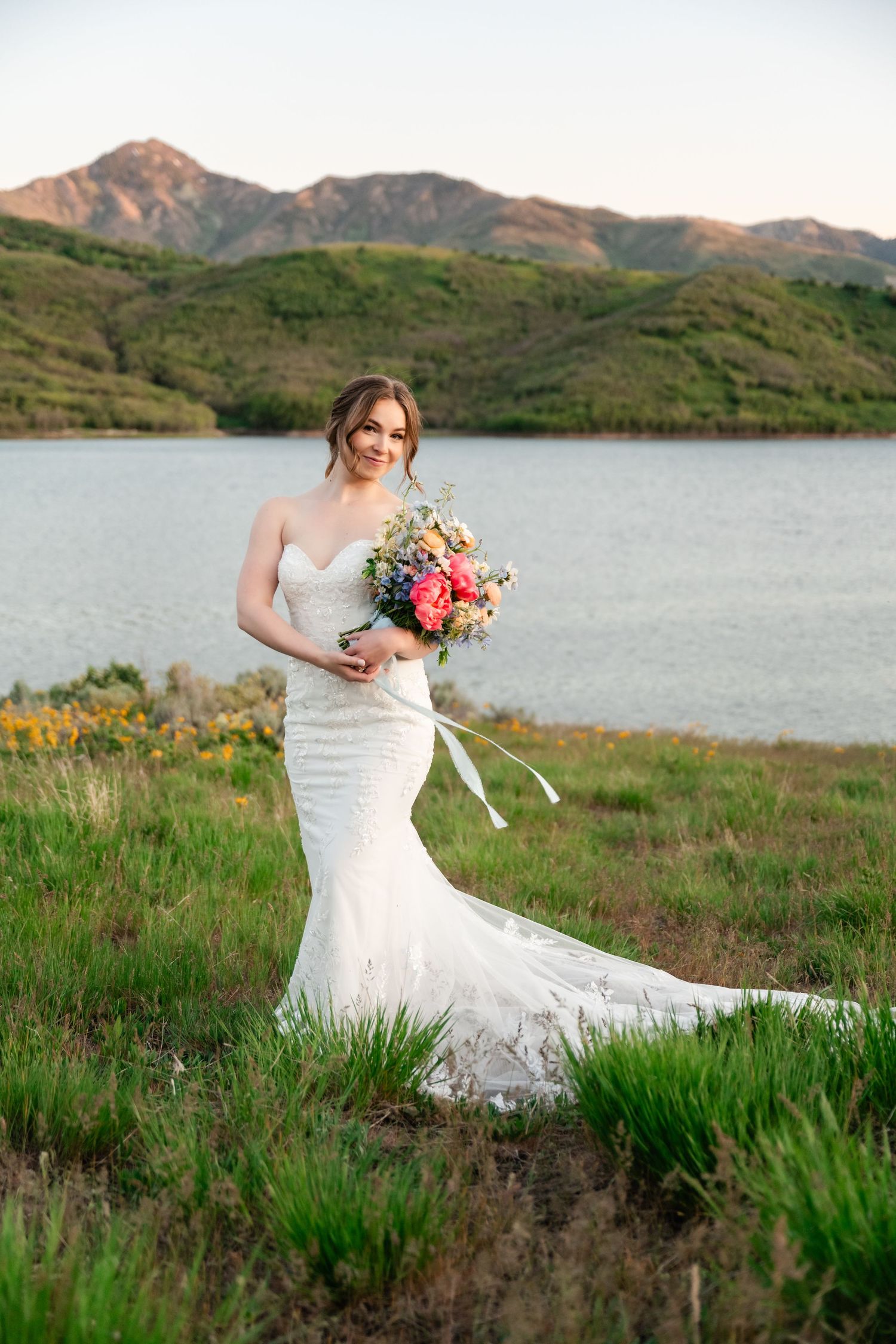 Bride in fitted lace gown holds colorful bouquet against mountain lake landscape.
