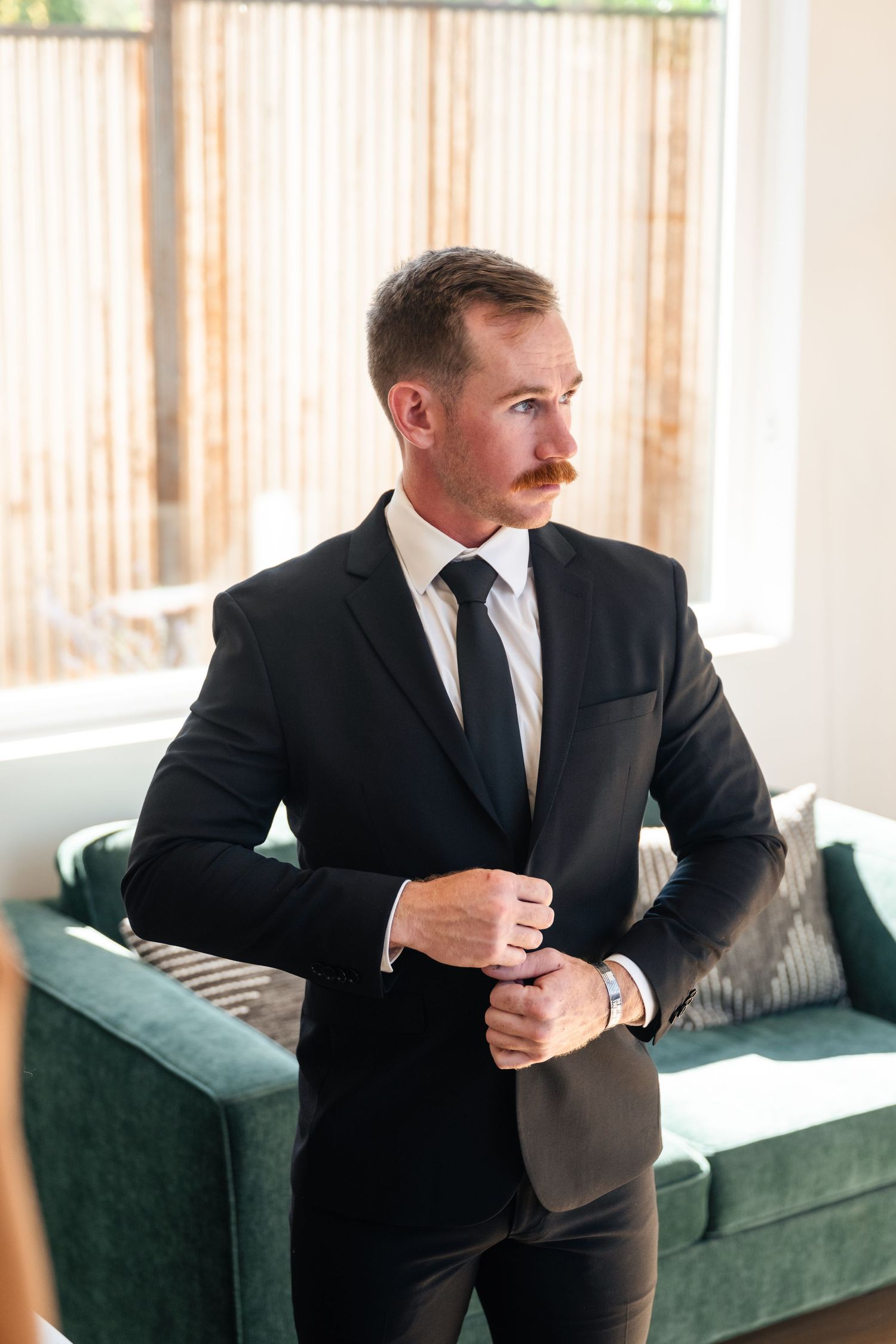 Person in black suit adjusts cufflinks while standing by green velvet couch.
