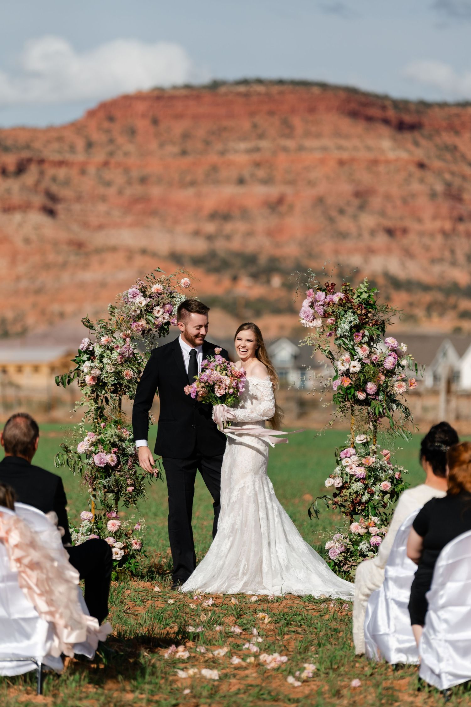 A wedding ceremony takes place against red rock cliffs with floral arches in an outdoor desert setting. Zion wedding photographer.