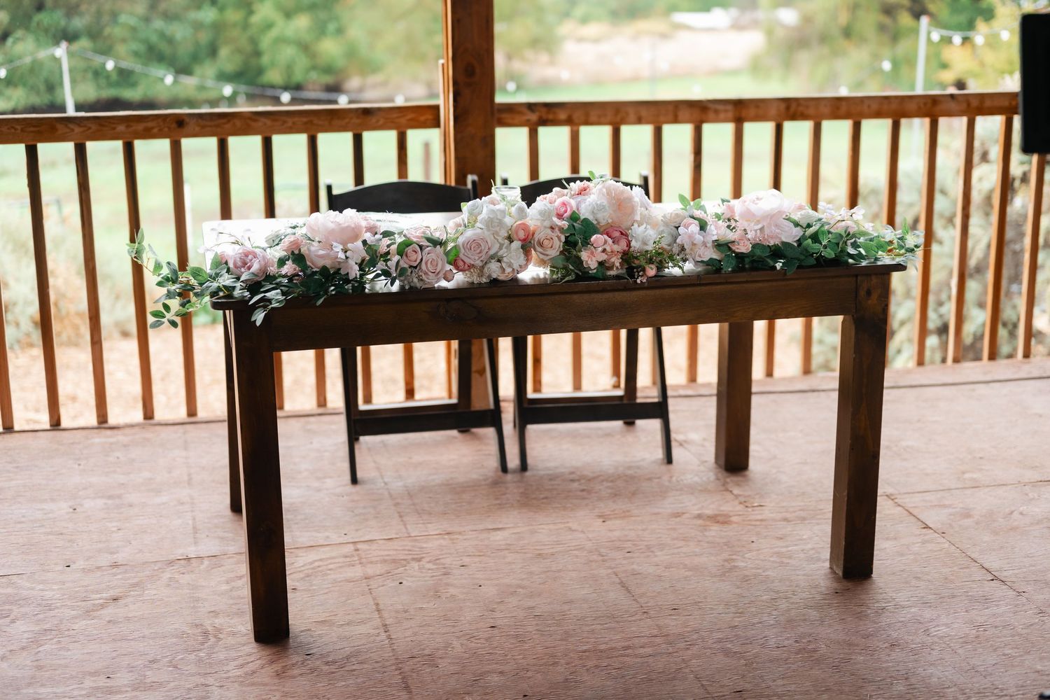 Wooden sweetheart table decorated with pink and white floral garland on covered porch.
