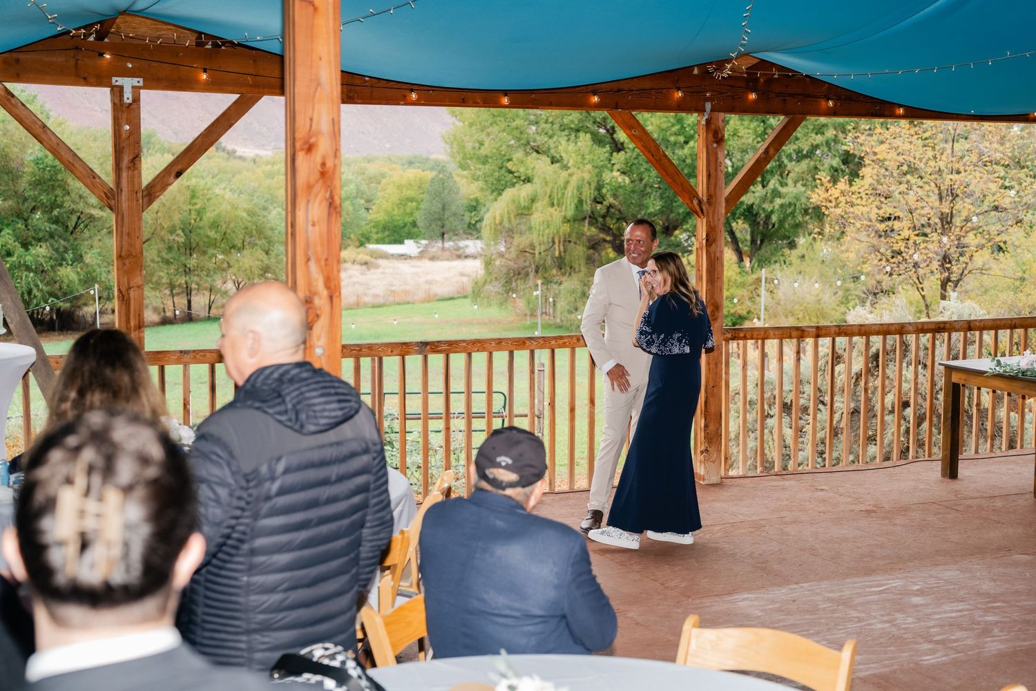 Guests gathered under a blue canopy on a wooden deck during a daytime event.