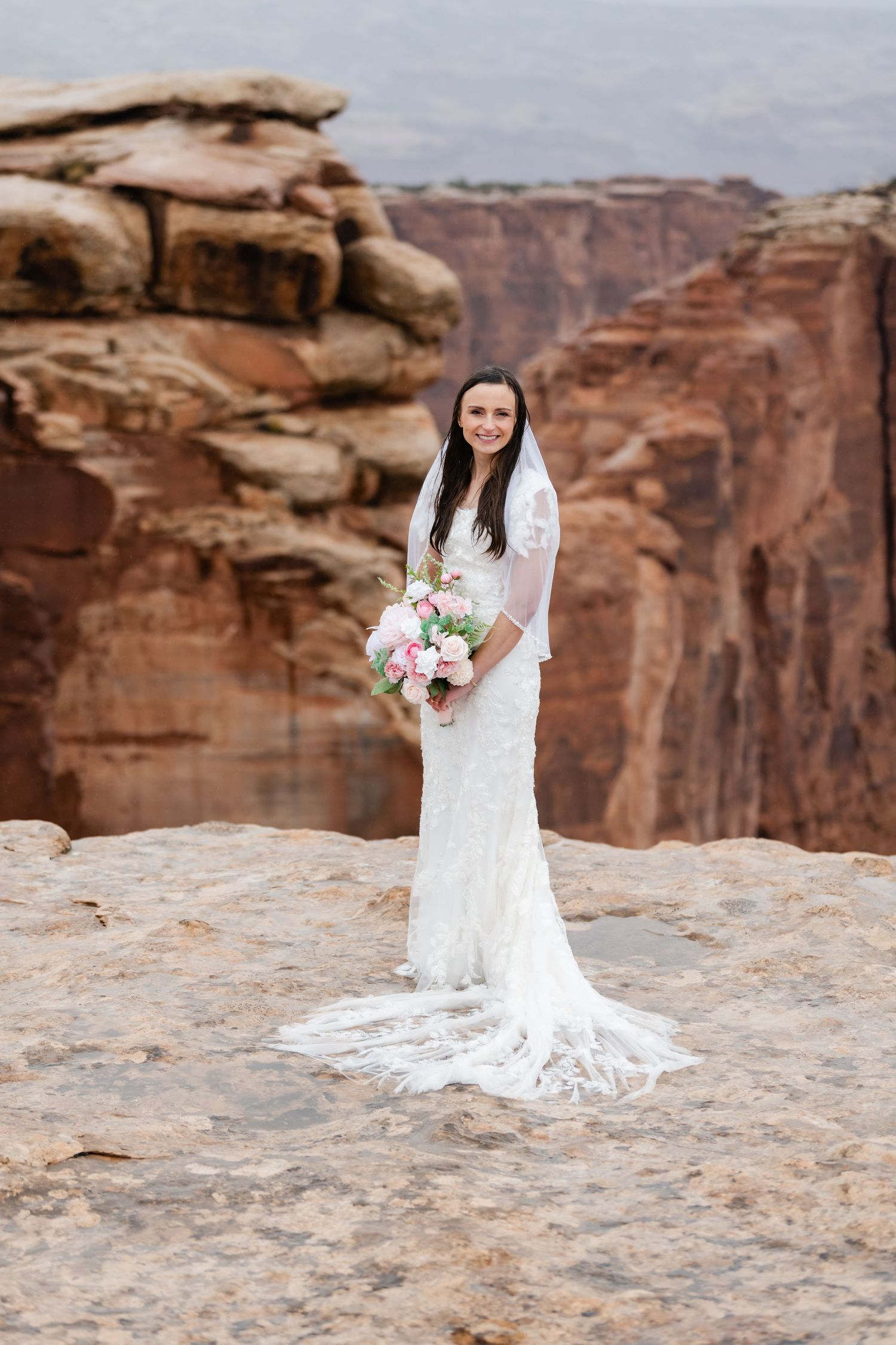 A bride in a white lace gown stands on red rock cliffs holding a pink and white bouquet.