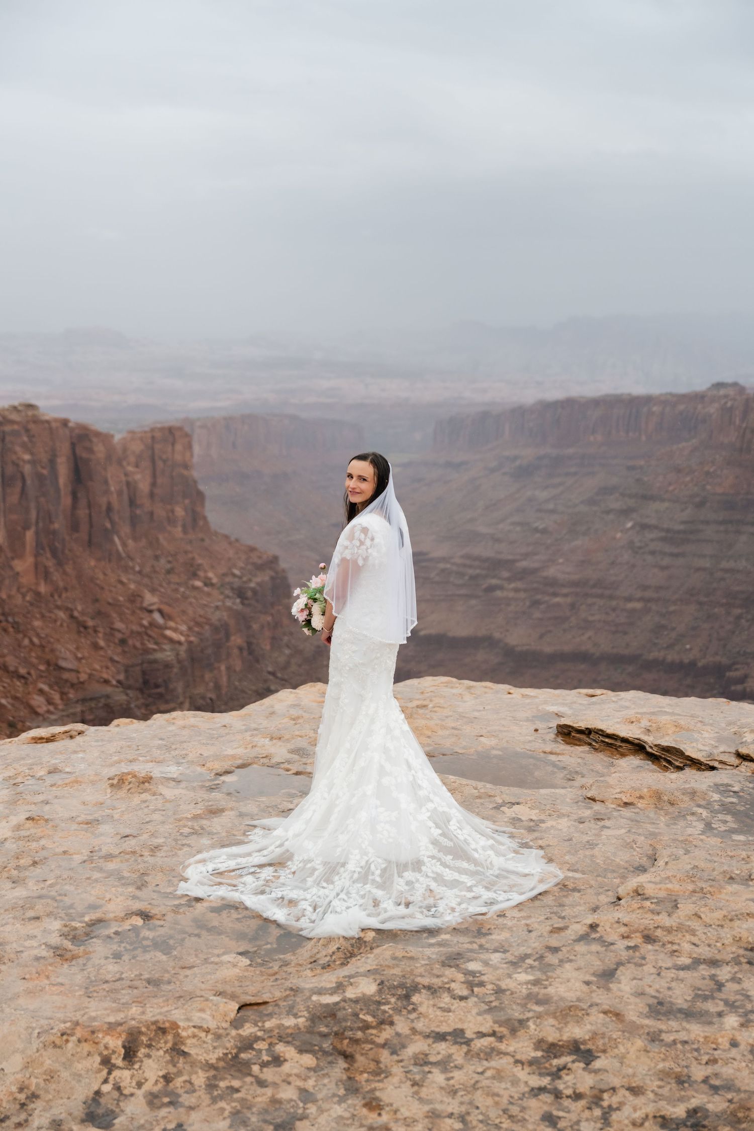 A bride in a flowing white dress with long train poses against a stunning canyon landscape.