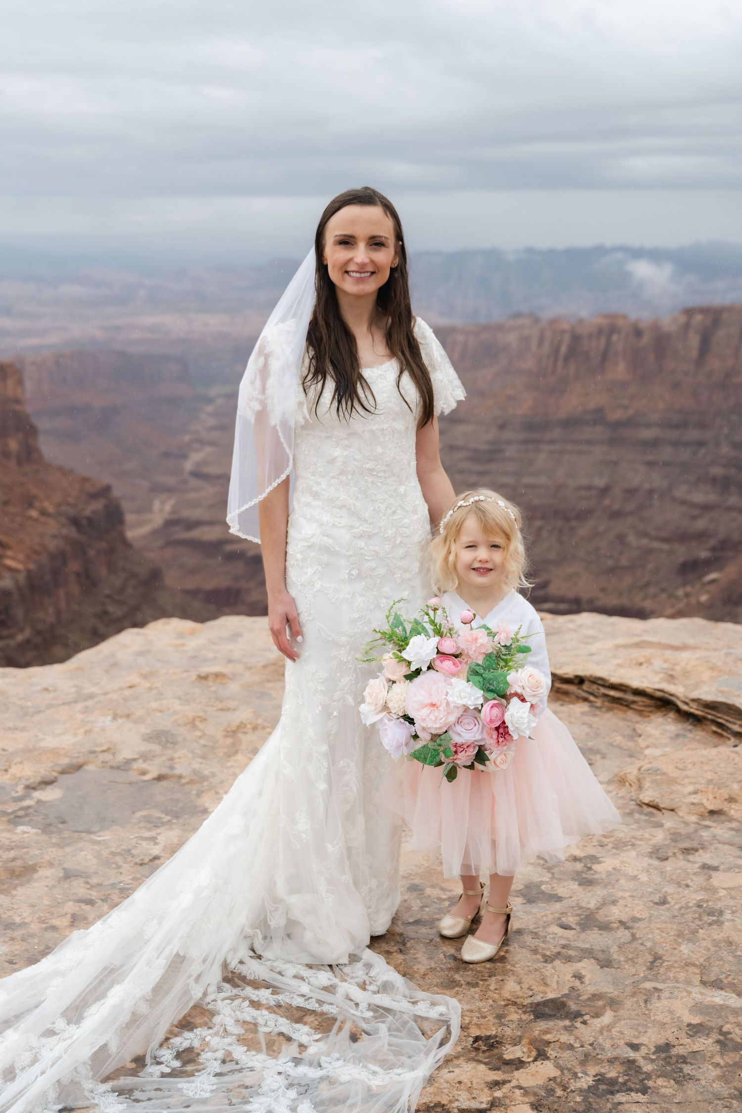 Bride in lace gown with flowing train poses with bouquet at canyon overlook.
