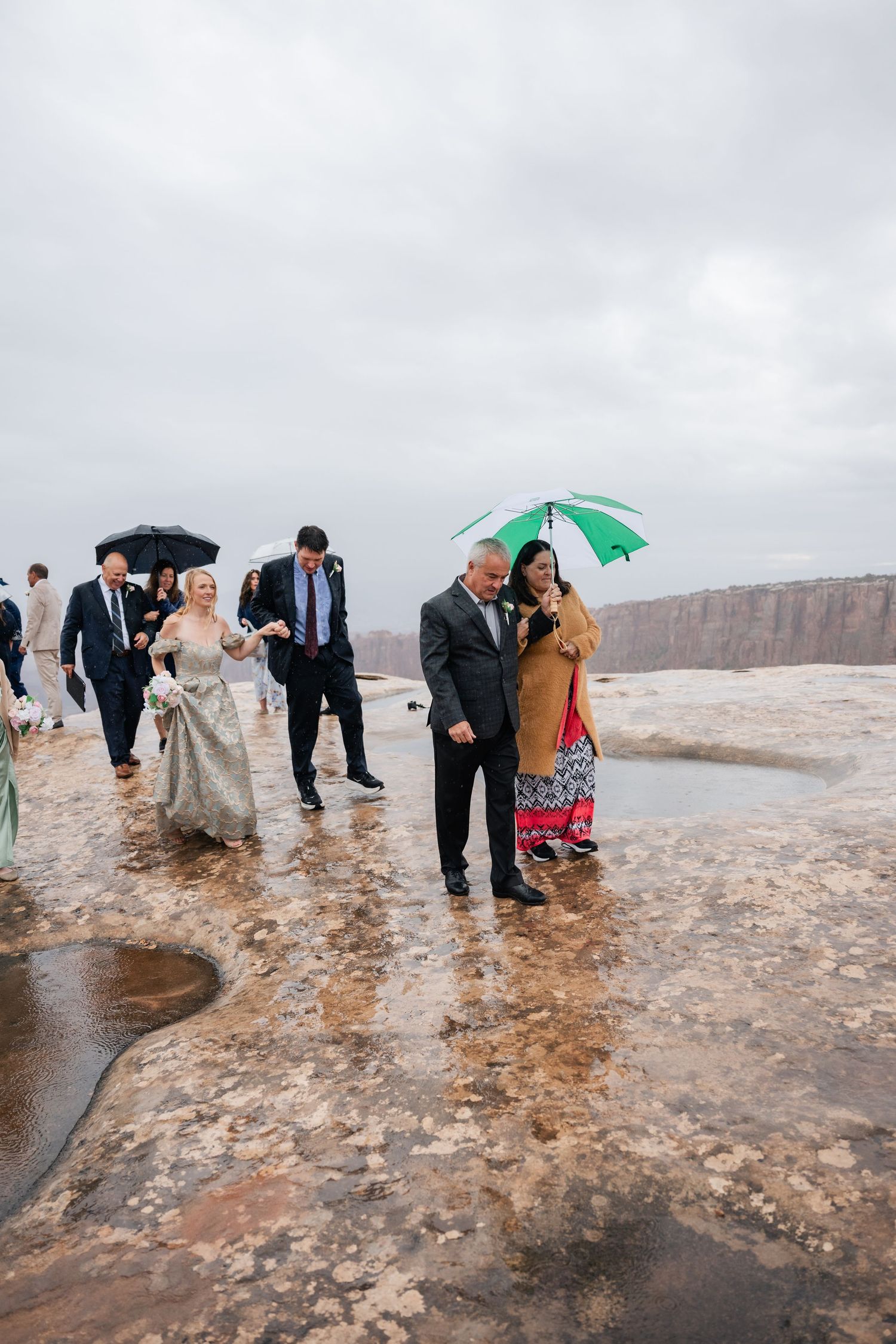 Wedding guests with umbrellas gather on wet rocky terrain during overcast weather.