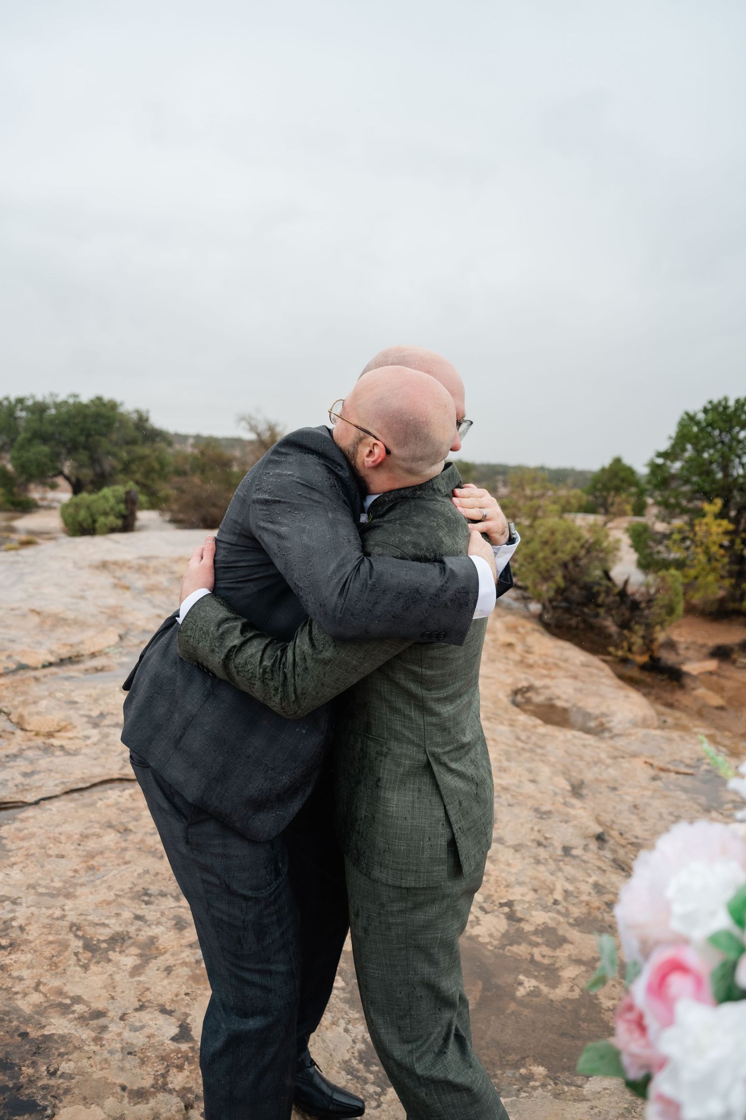 Two people sharing an emotional embrace on a rocky desert overlook with flowers in the foreground.