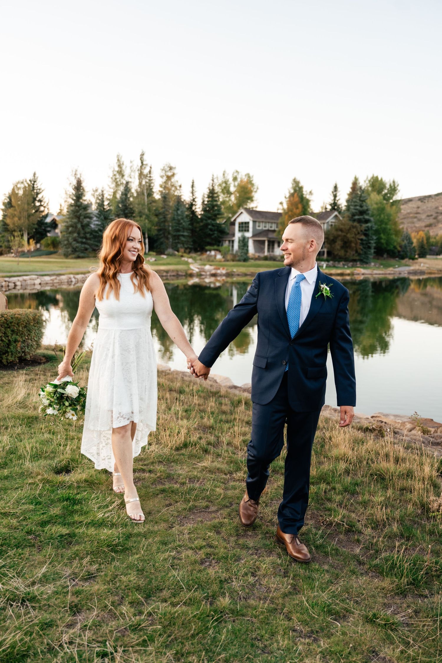 A couple walks hand in hand through tall grass by a lake, with the bride in white and groom in navy suit.