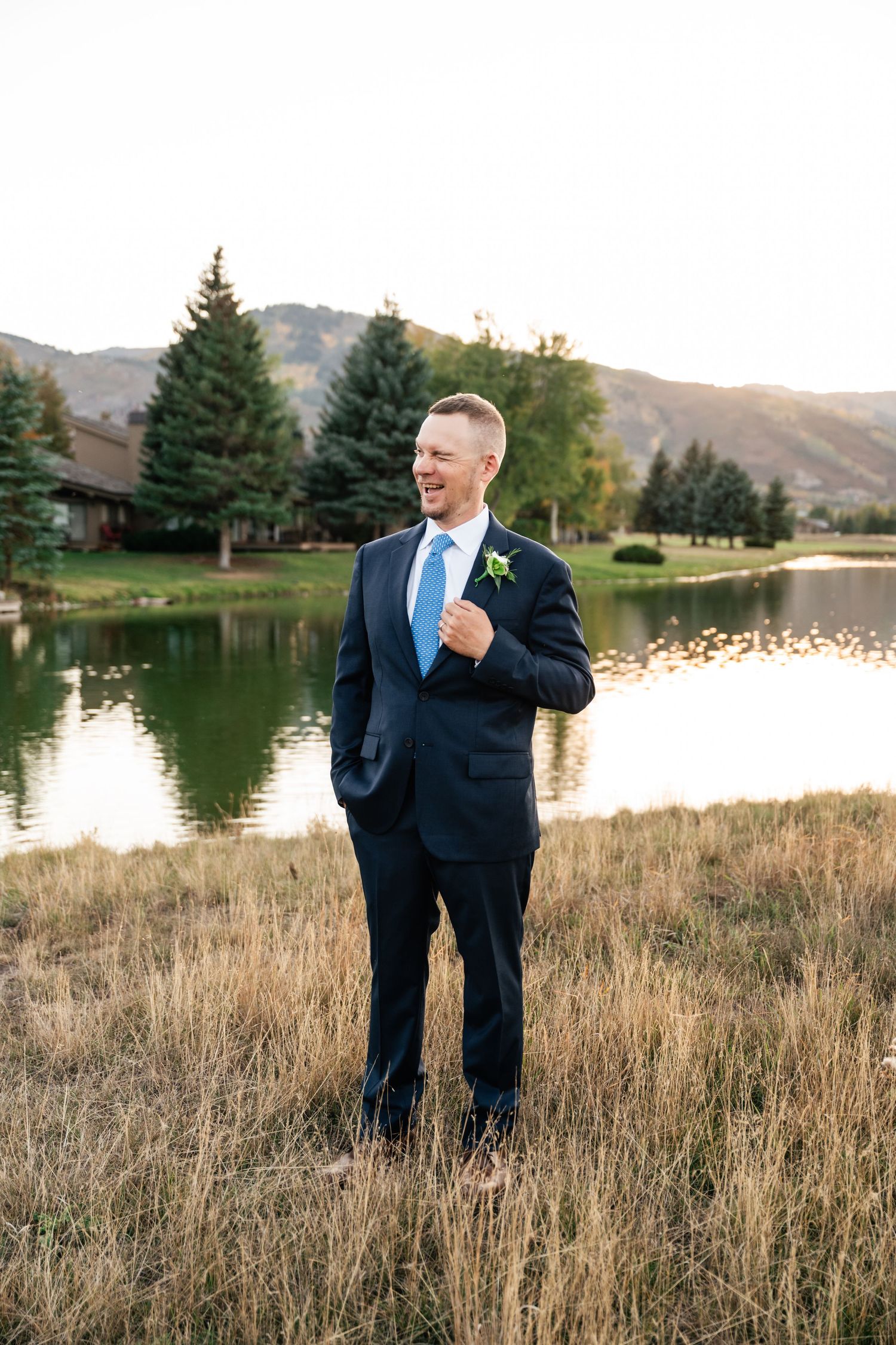 A person in a navy suit stands in tall grass near a tranquil lake with mountains and evergreen trees in the background.
