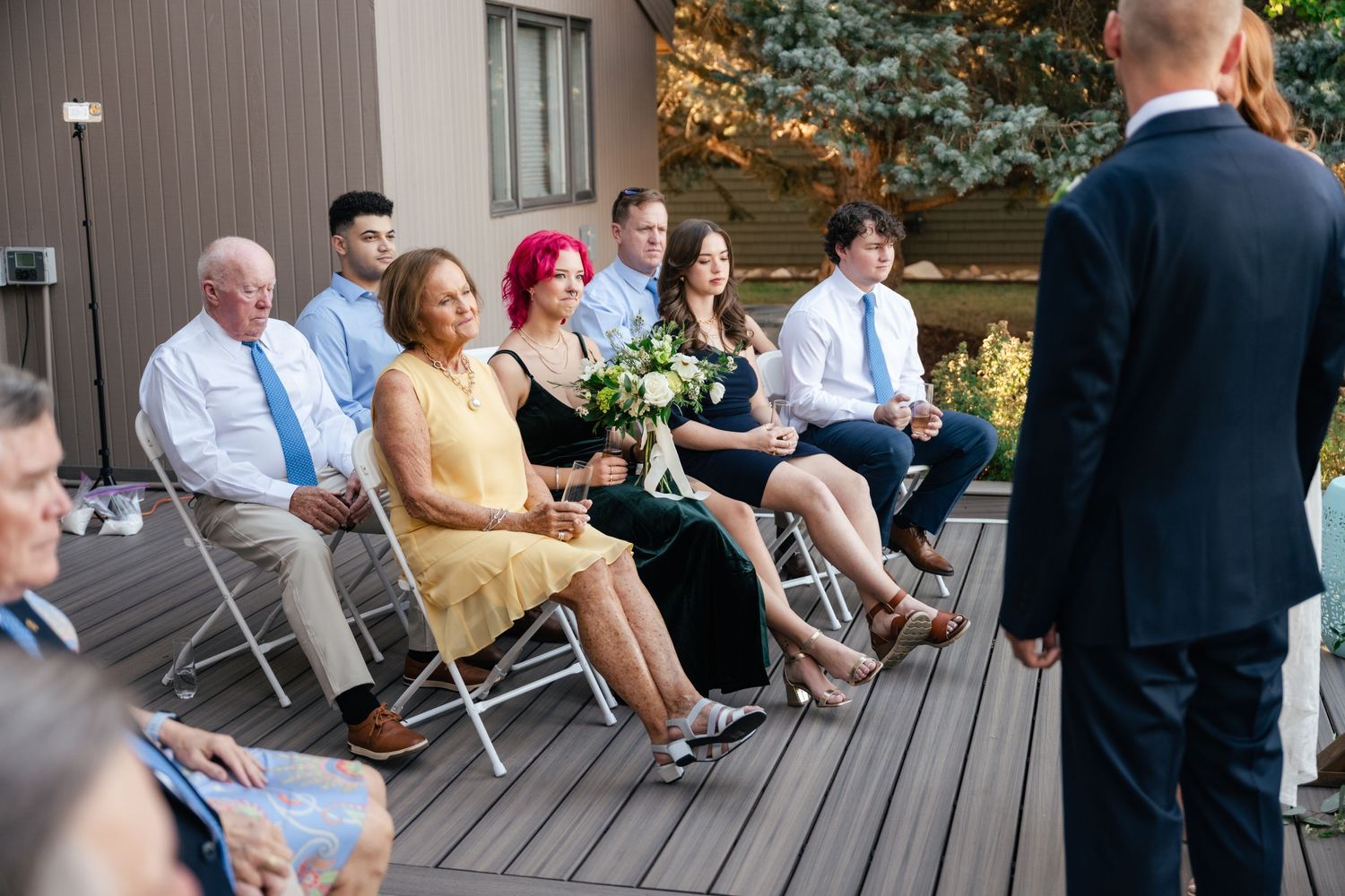 Wedding guests seated in folding chairs on a wooden deck during an outdoor ceremony.
