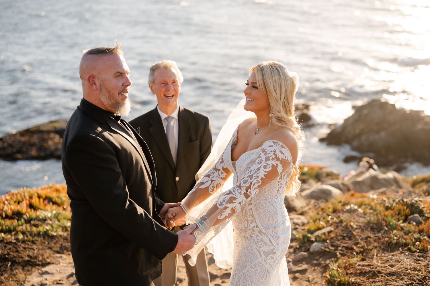 An intimate outdoor wedding ceremony unfolds on a rocky oceanside cliff bathed in warm sunlight.