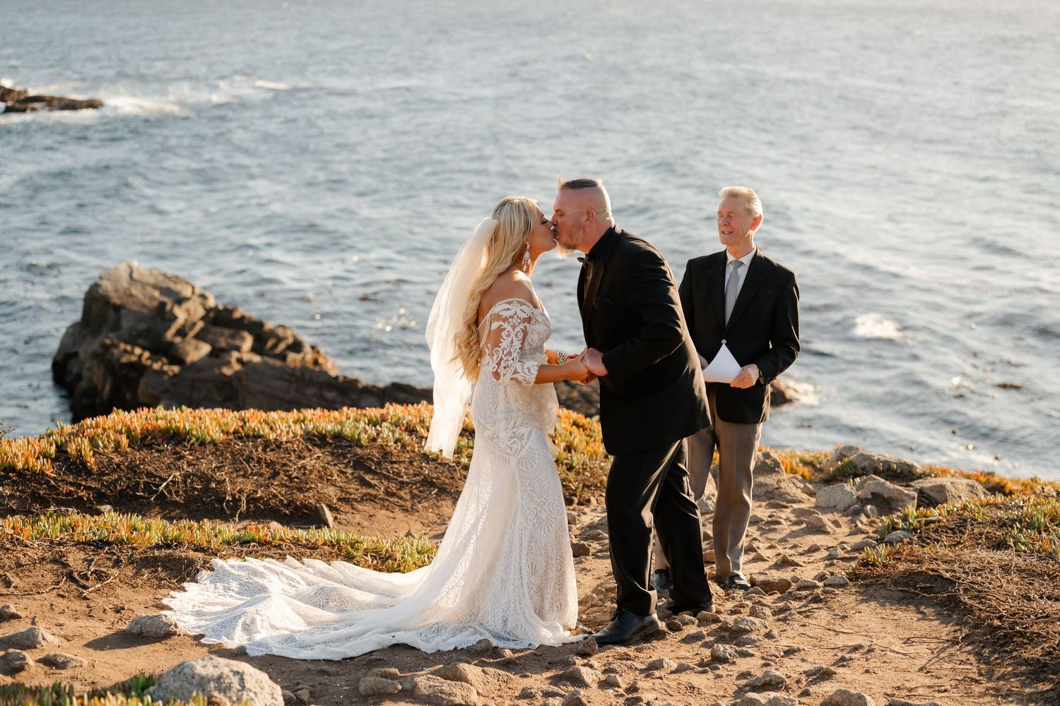 Intimate coastal wedding ceremony taking place on rocky cliffs overlooking the ocean at sunset.