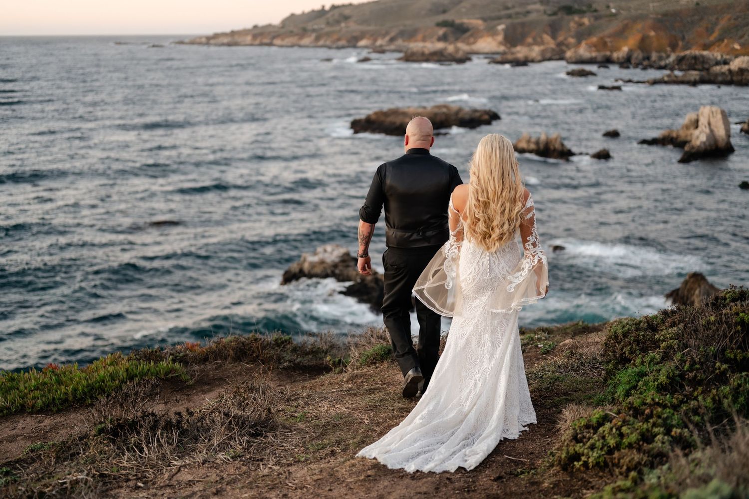 A wedding couple stands on dramatic coastal cliffs overlooking the Pacific Ocean at golden hour.