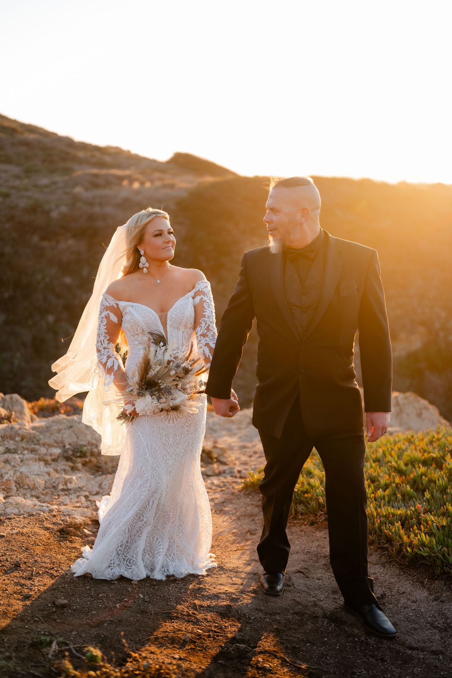 A couple in wedding attire walks along a scenic hilltop path at golden hour.