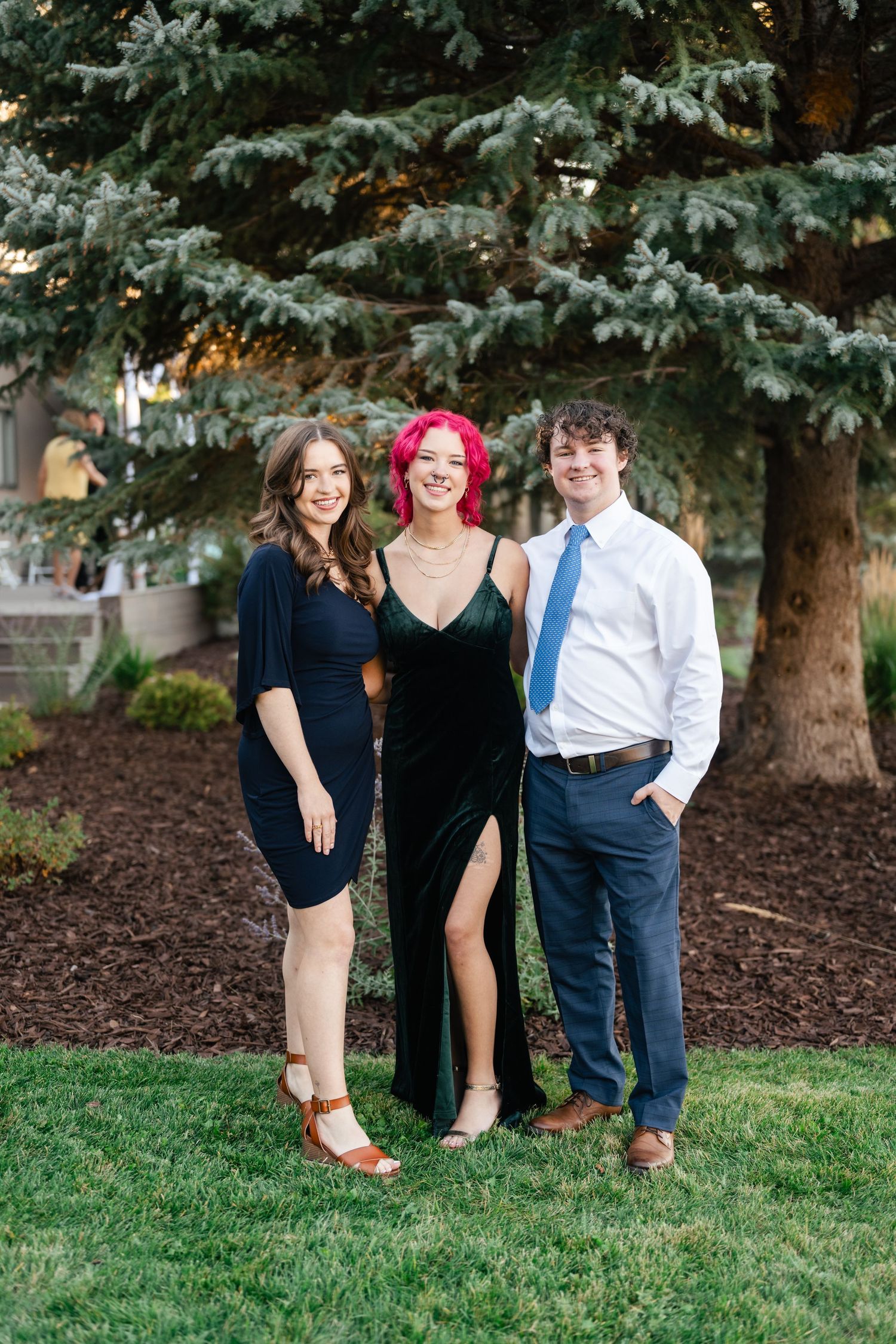 Friends pose playfully together on grass near evergreen trees.