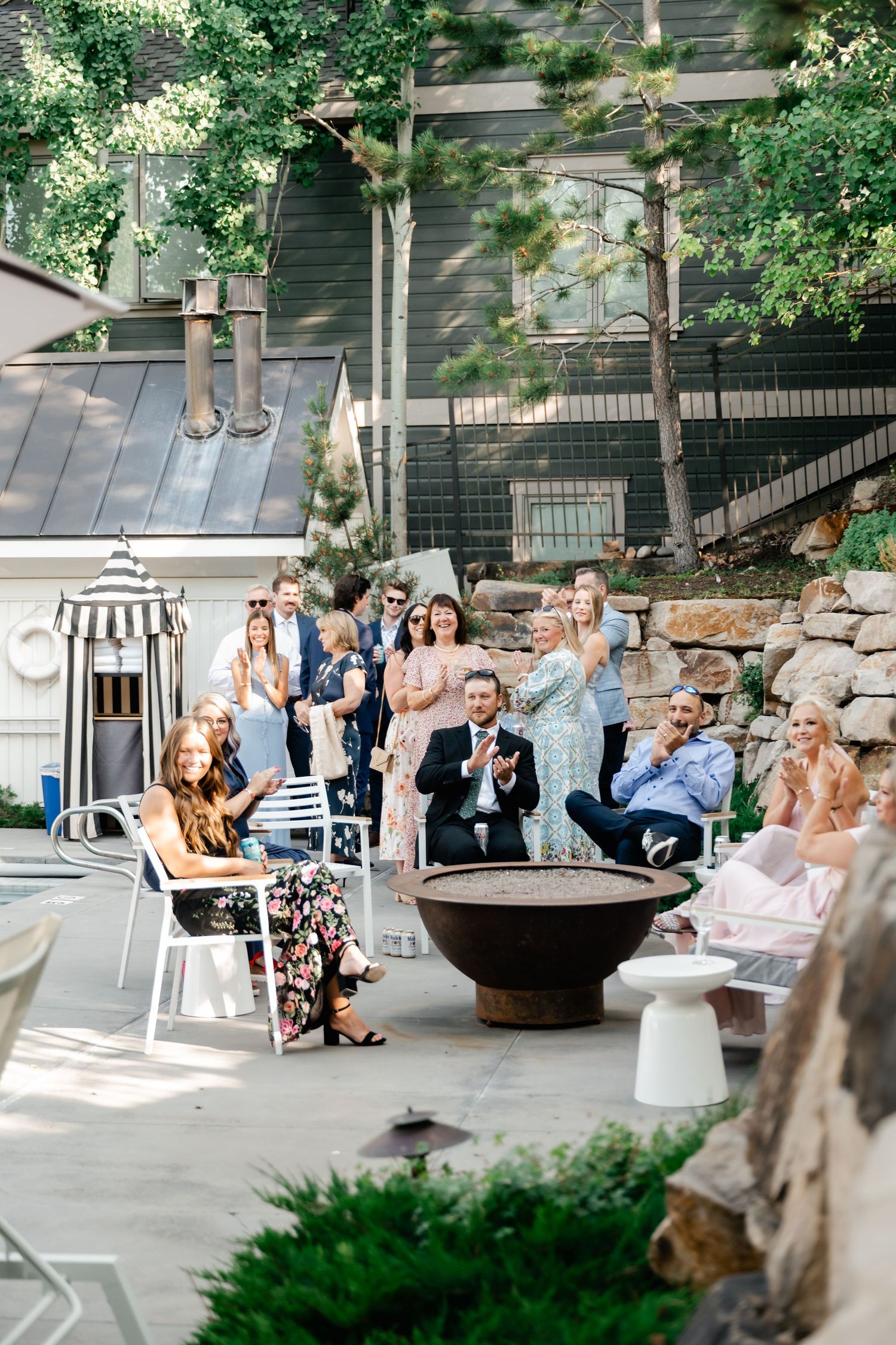 Wedding guests seated outdoors on white chairs celebrate during a garden ceremony.