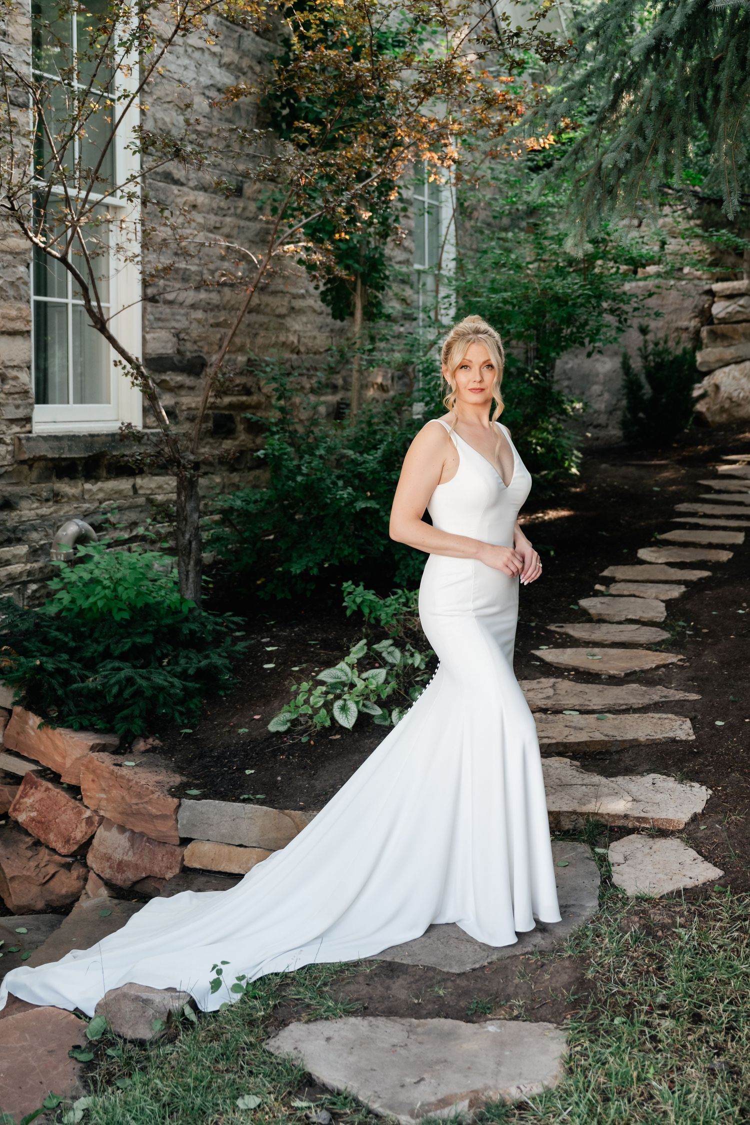 A stunning mermaid-style wedding dress is showcased against a rustic stone wall backdrop with a winding garden path.
