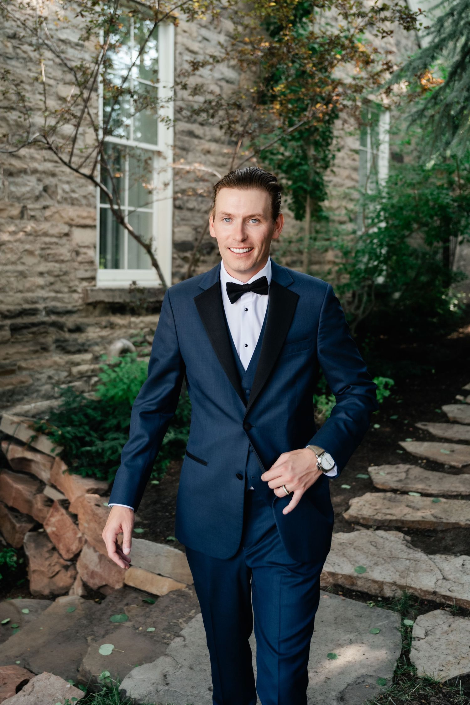 Groom in navy tuxedo poses against stone wall backdrop with natural lighting.
