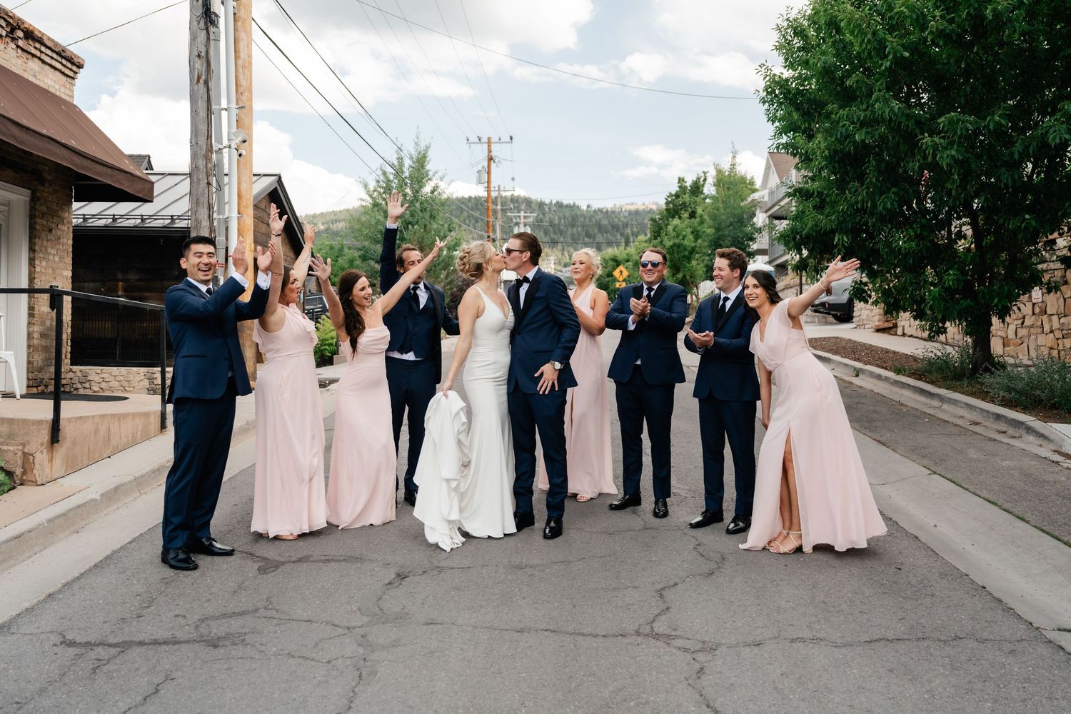 Wedding party in formal attire walking along a street with trees and buildings visible.