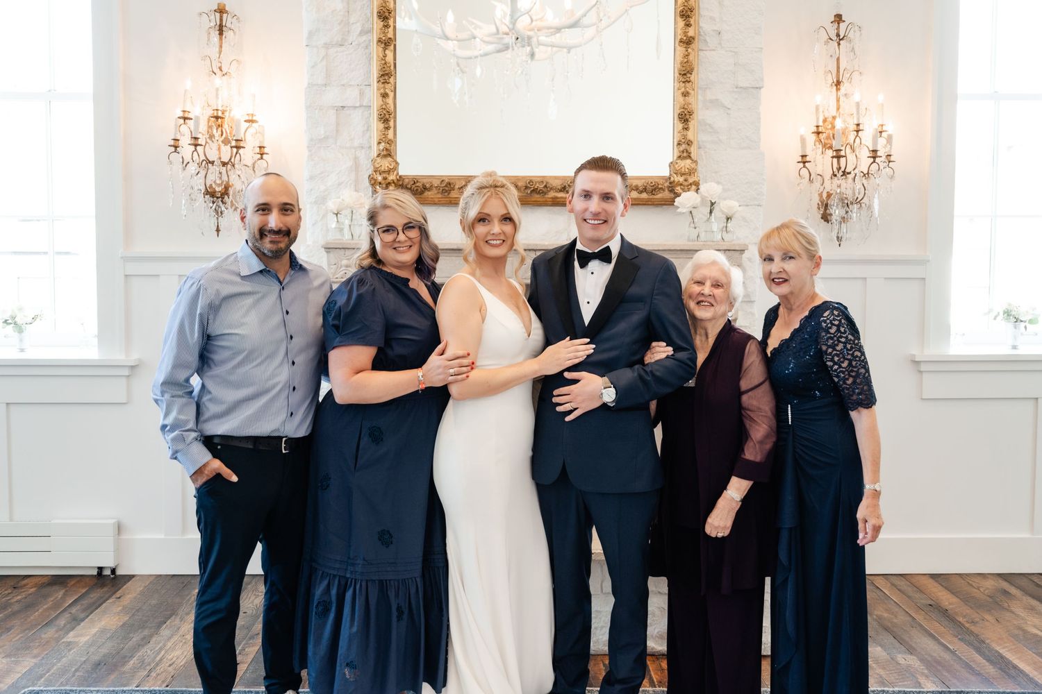 A group in formal attire poses together in an elegant room with crystal chandeliers and white walls.