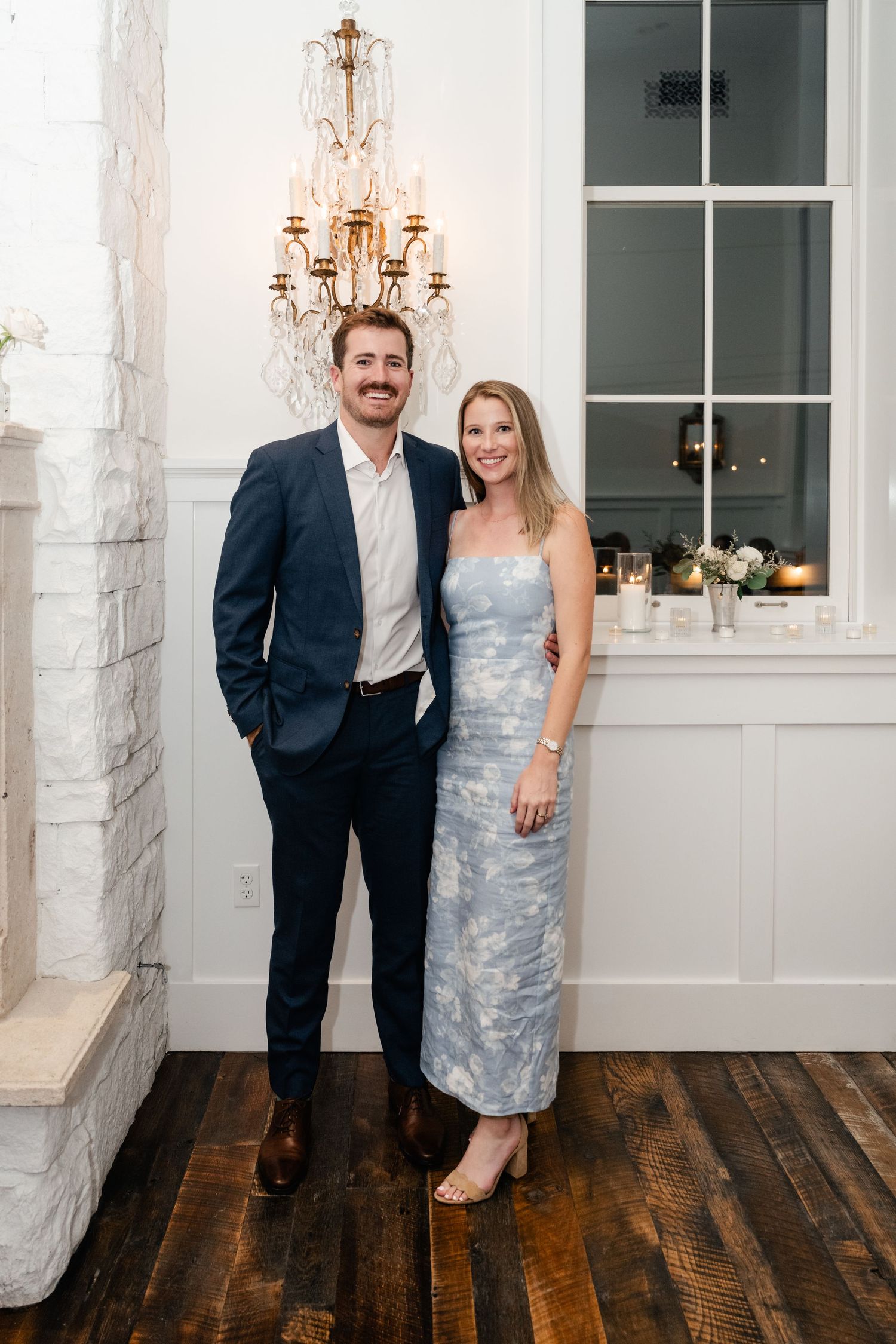 A couple poses together in formal attire against a white wall with elegant lighting fixtures.