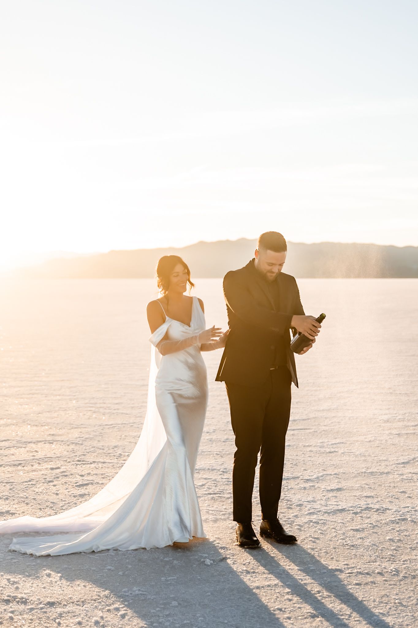 Newlyweds in formal attire share candid moments on vast salt flats during sunset.