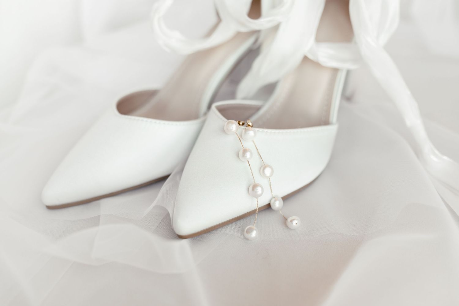 Elegant white satin wedding shoes with pearl embellishments displayed on delicate white tulle fabric.