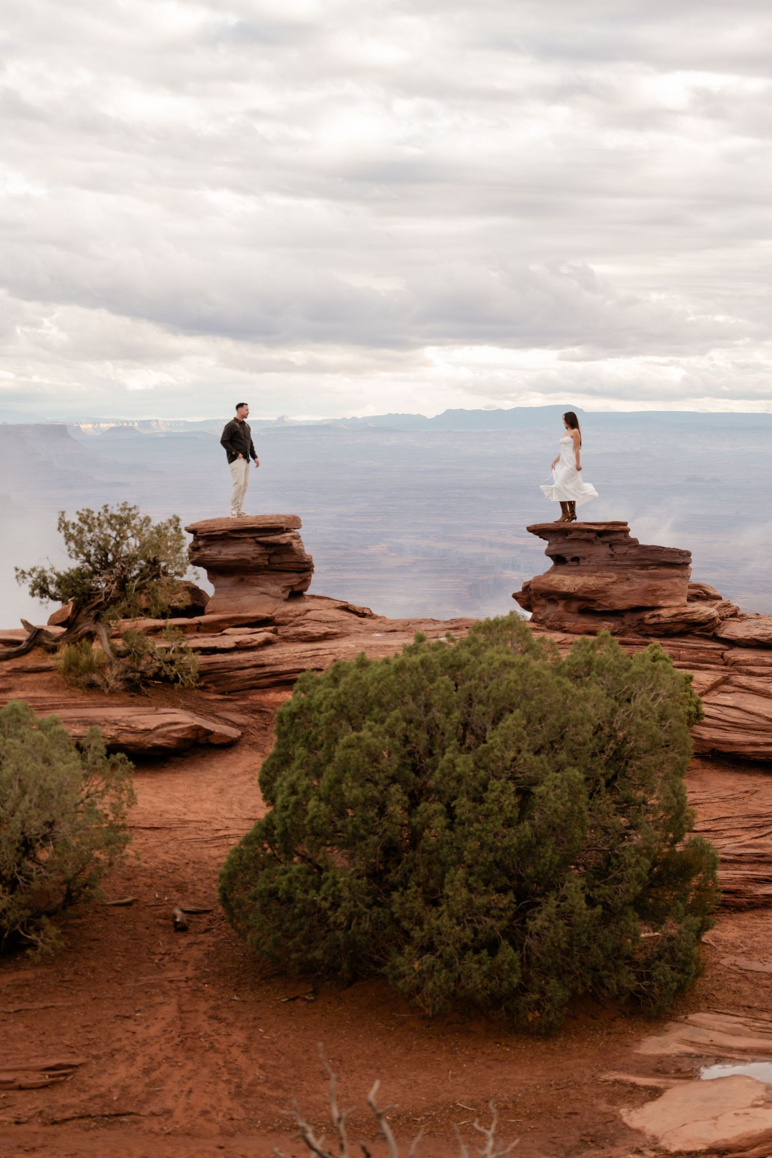 Two people stand on separate red rock formations in a dramatic desert landscape with cloudy skies and distant mountains.