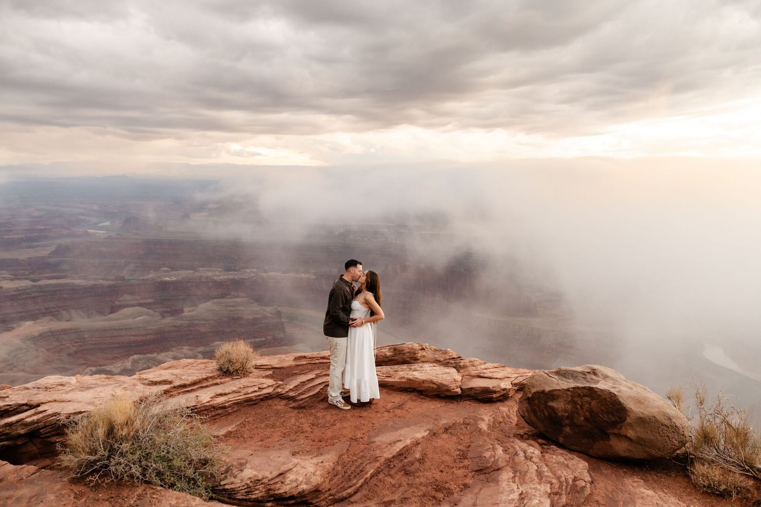 Couple embracing on red rock overlook during misty sunset with vast canyon views.