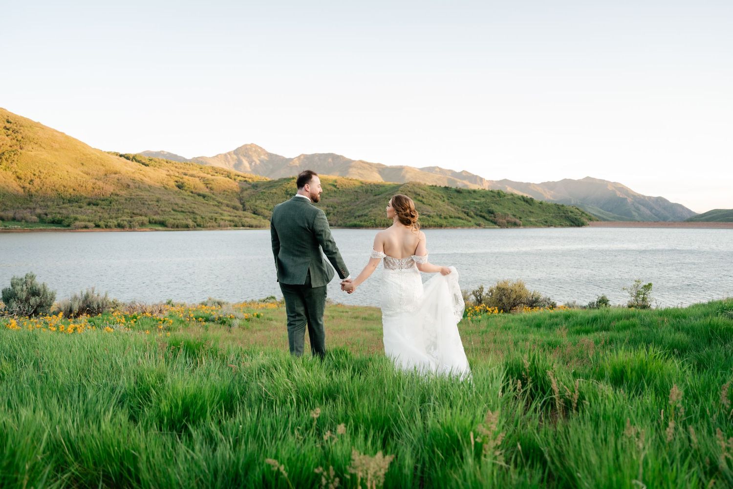 Panoramic view of wedding couple walking through green meadow near mountain lake.