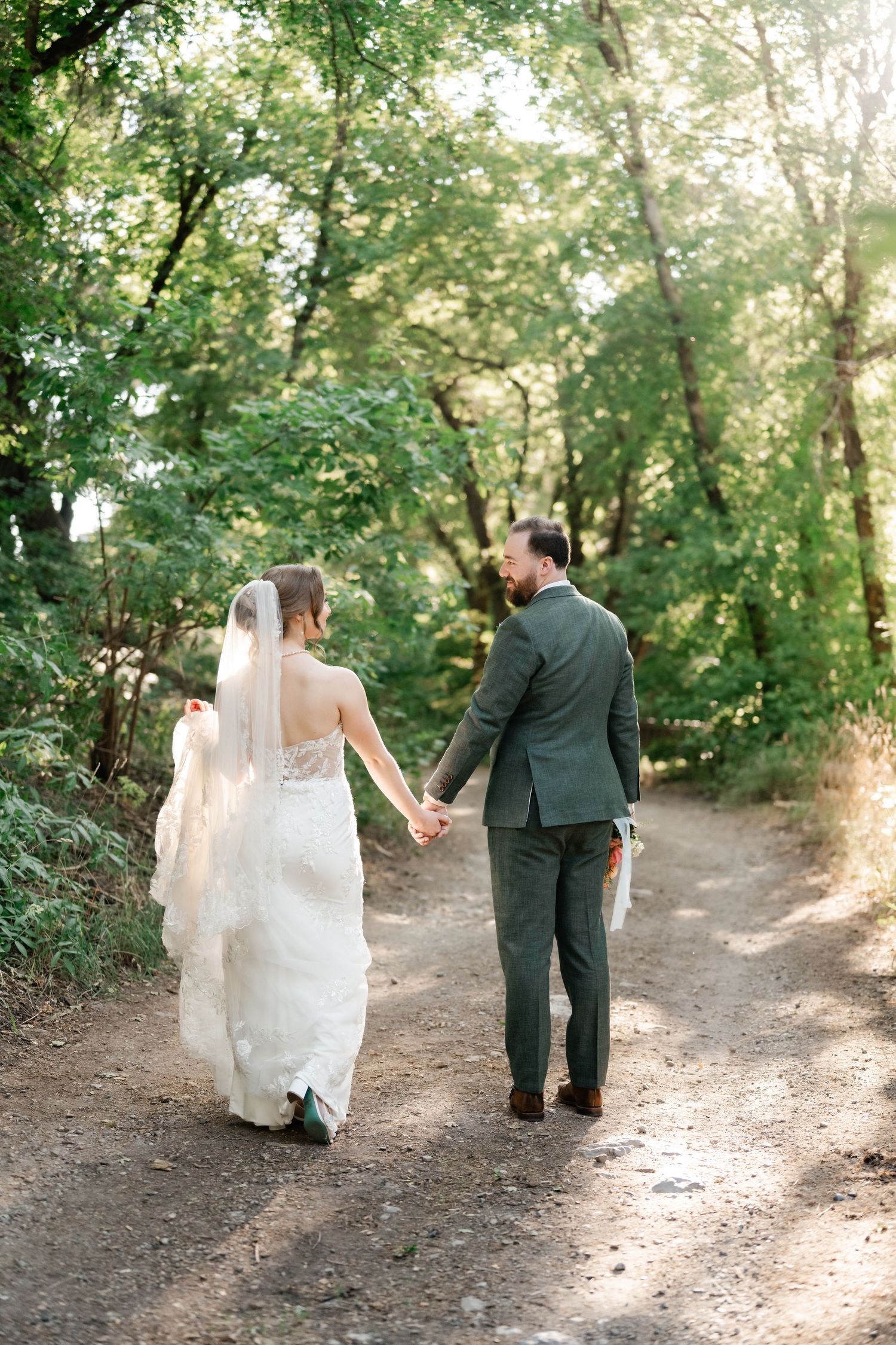 A couple walks hand in hand down a sunlit forest path during their wedding photoshoot. Millcreek Inn Wedding Photographer.