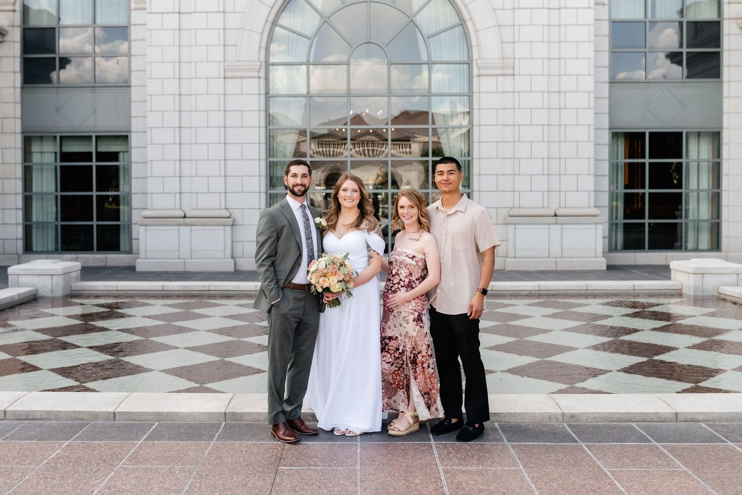 Wedding party members pose together against a white stone building with arched windows.