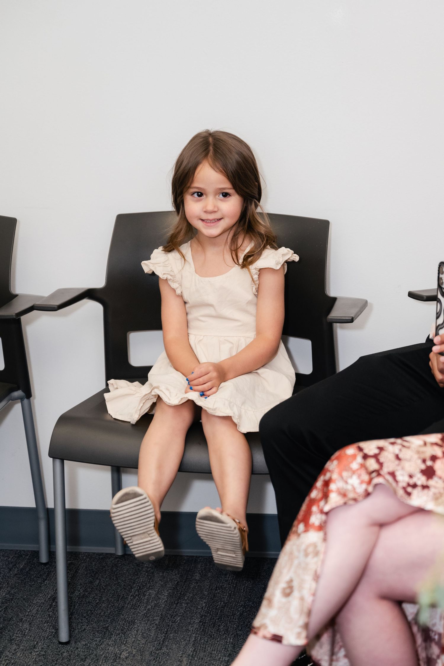 A young child in a cream-colored dress sits on a black chair in what appears to be a waiting room.