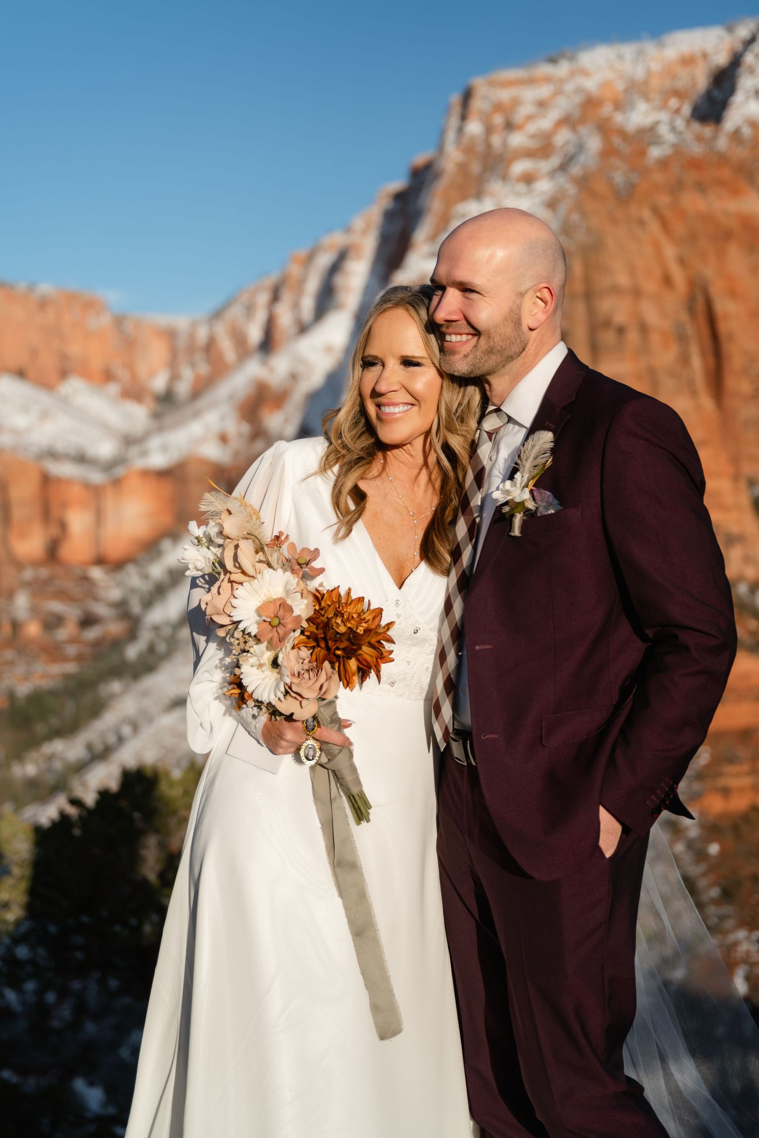 Couple in white dress and burgundy suit embrace while holding dried flower bouquet against mountain backdrop.