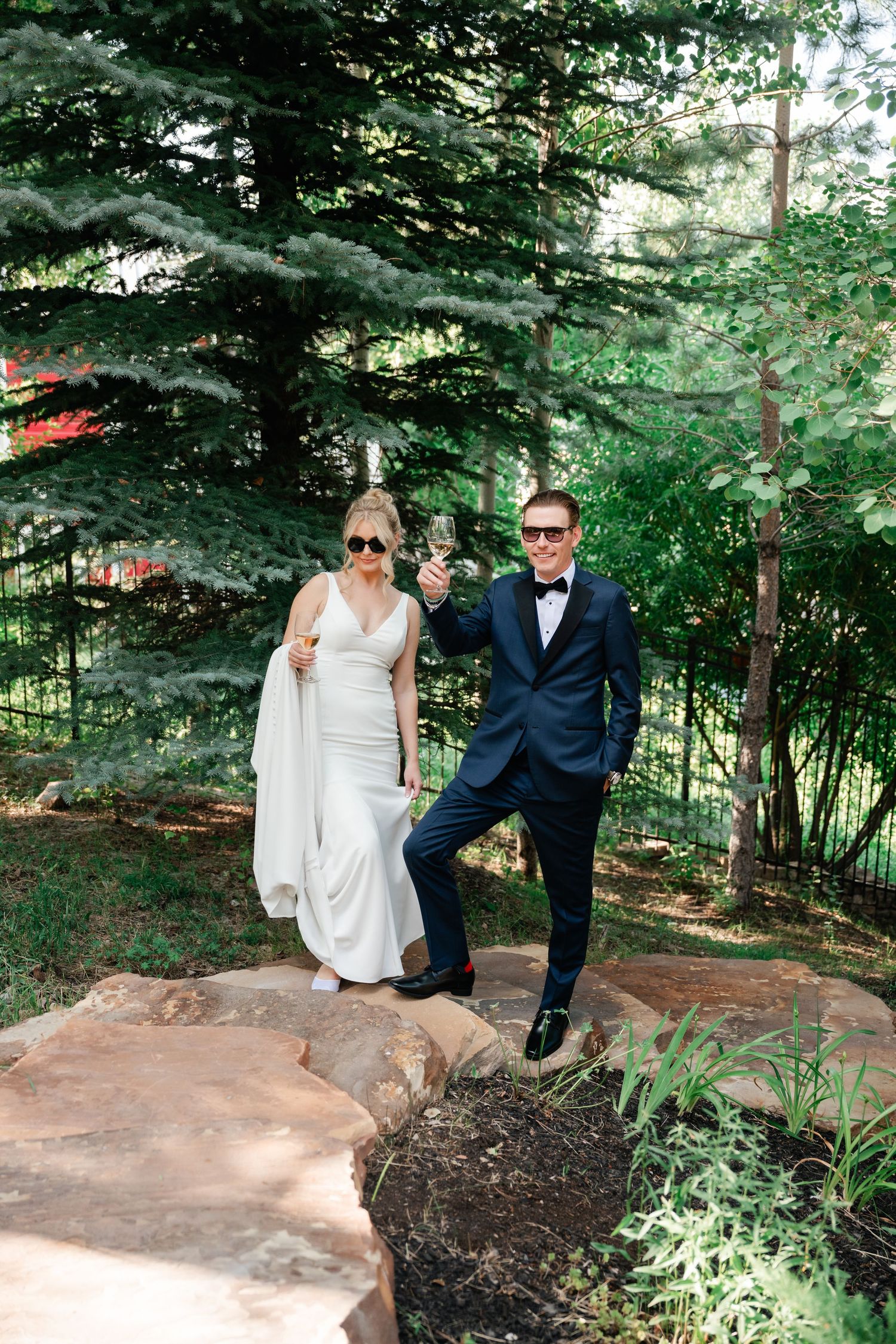 A couple in wedding attire poses playfully on a large rock in a wooded garden setting. Chill wedding couple at their Park City Wedding.