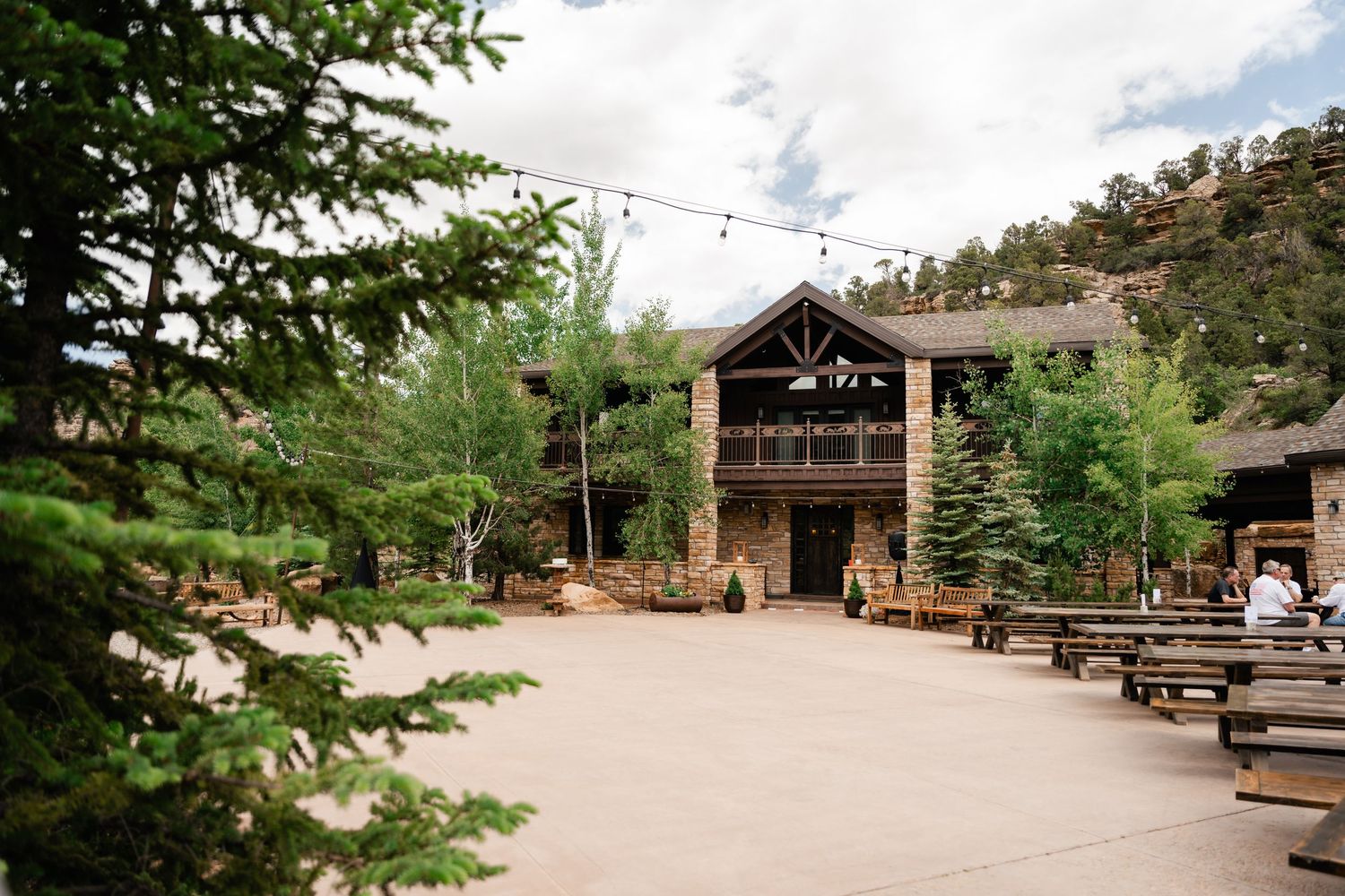 Rustic wooden picnic pavilion with log construction and stone foundation nestled among pine trees in a mountain setting.