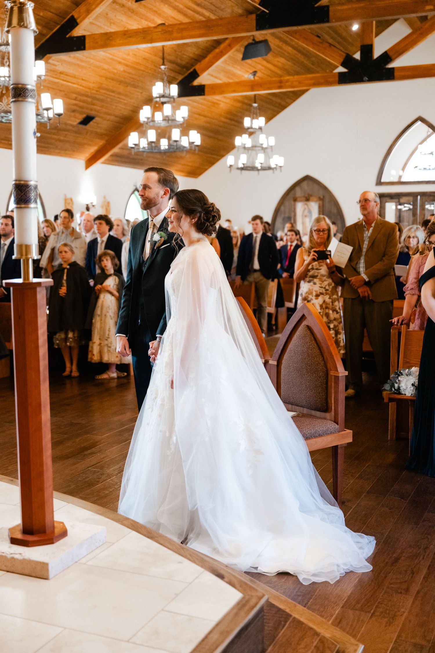 Wedding ceremony in a rustic wooden church hall with elegant chandeliers and wooden beams overhead.