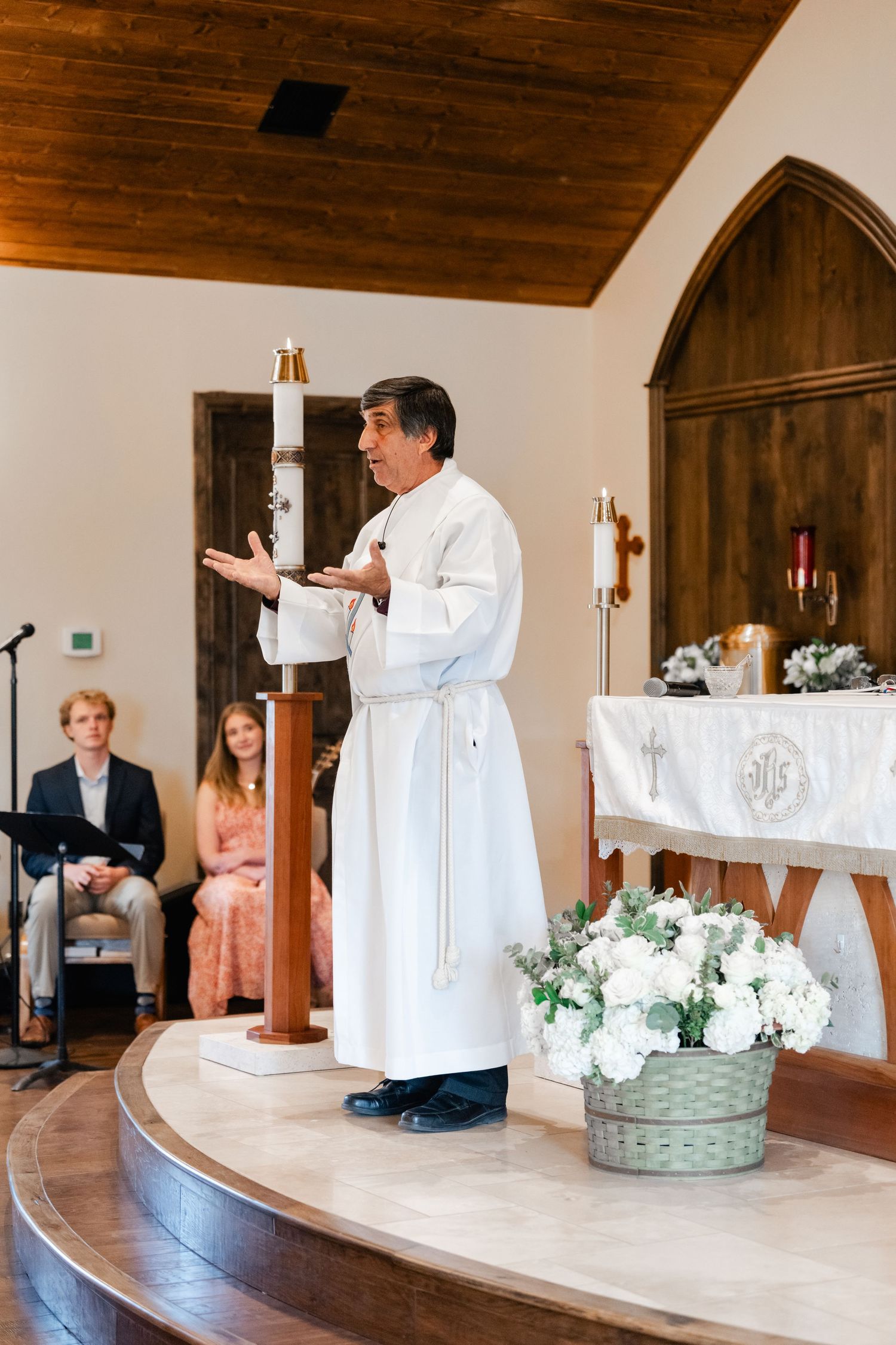 Priest in white robes conducts ceremony at altar decorated with flowers in traditional church setting.
