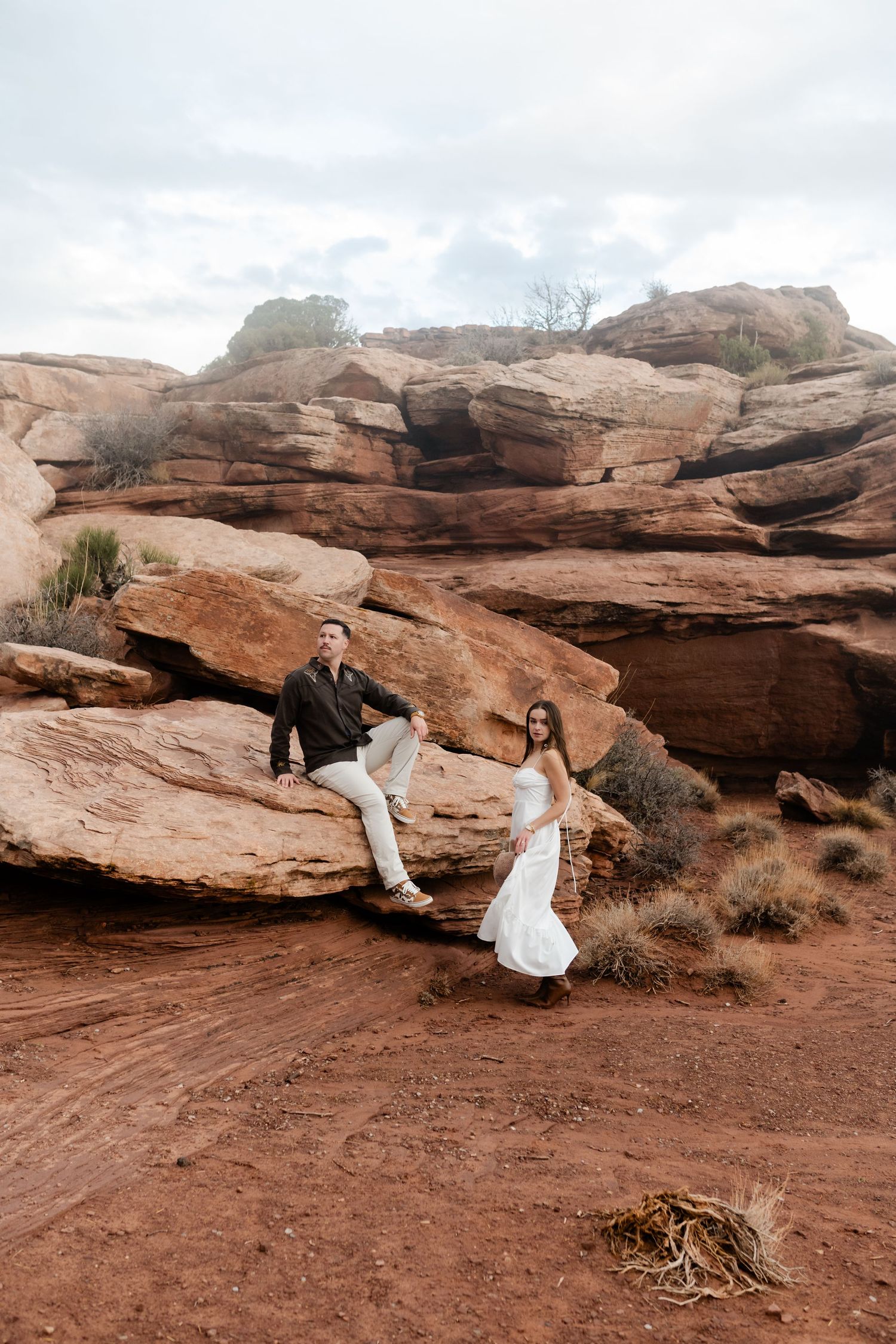 A couple poses playfully on red rock formations in a desert landscape during what appears to be an engagement photoshoot.