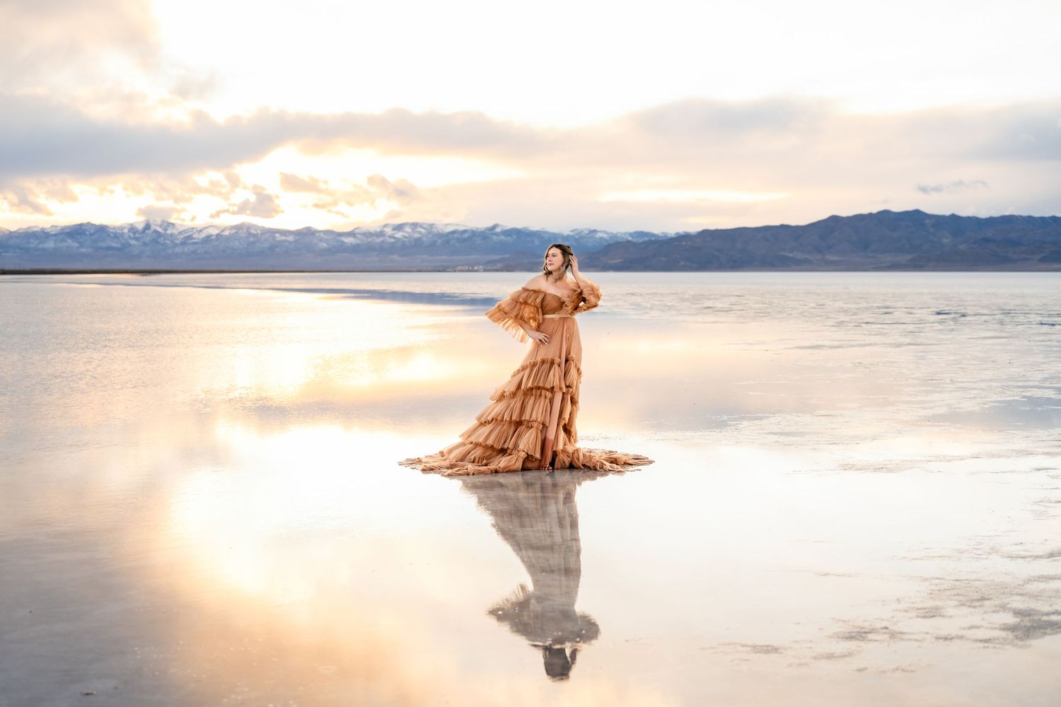 A flowing peach dress reflects in still water at sunset with snow-capped mountains in the background. Utah Salt Flats Photographer.