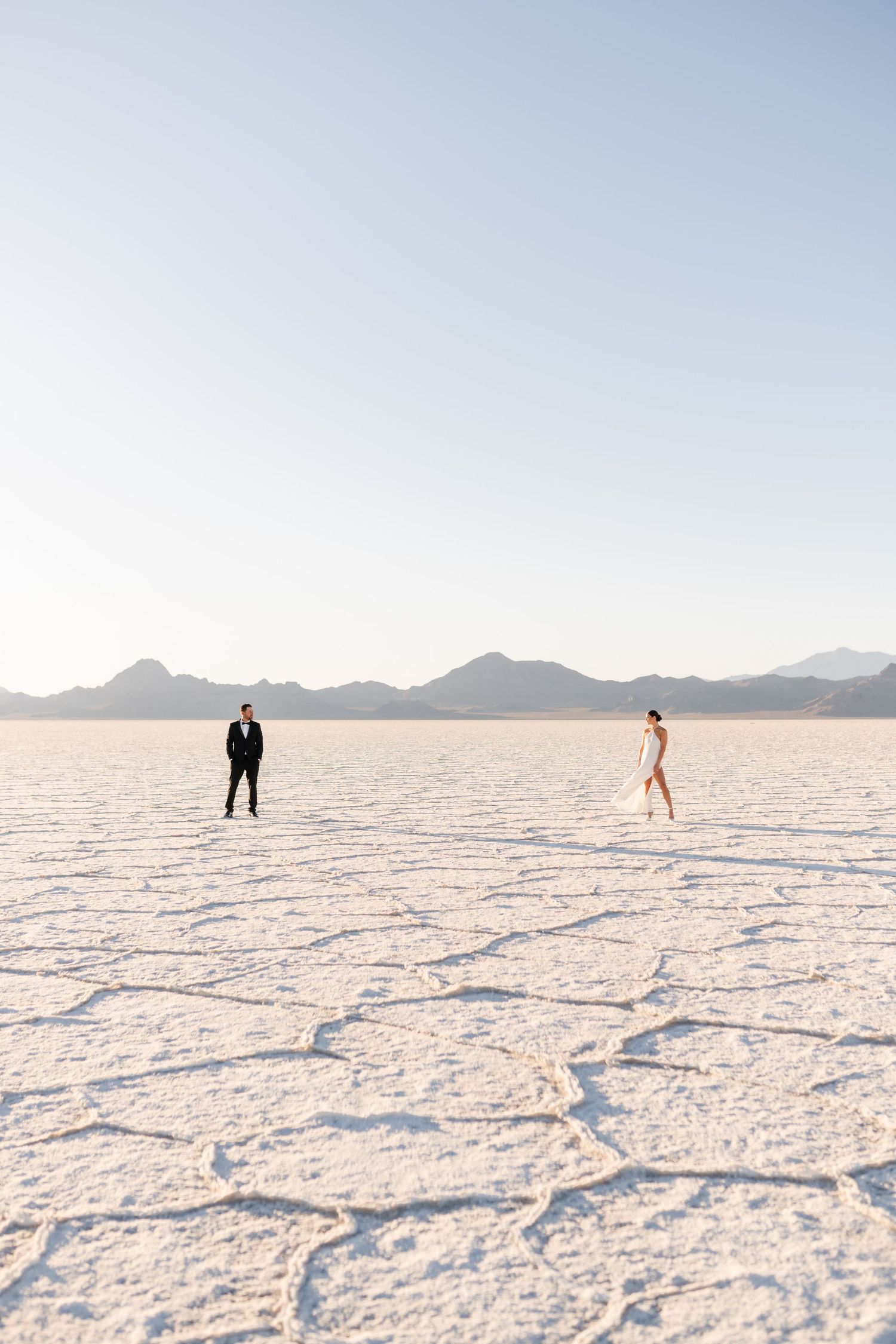 Salt Flats Photographer. Two figures standing far apart on a vast white salt flat with mountains in the distance during daytime.