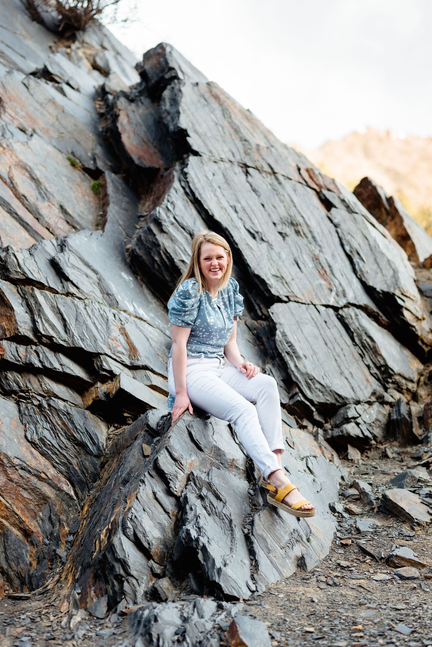 Person in blue shirt and white pants sits casually on large jagged rock formations during outdoor photo session.